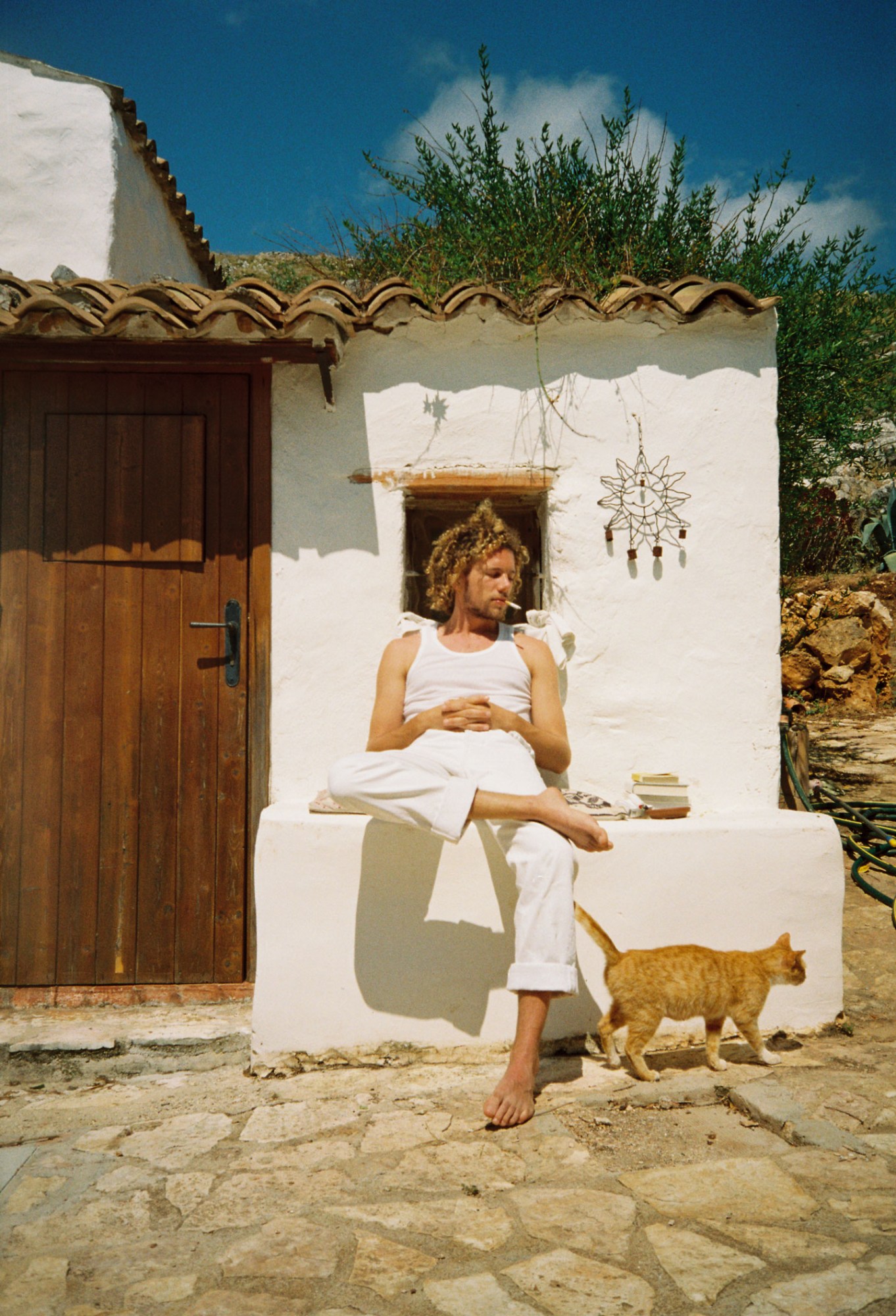 a long-haired man sits in front of a white Mediterranean-looking house with an orange cat