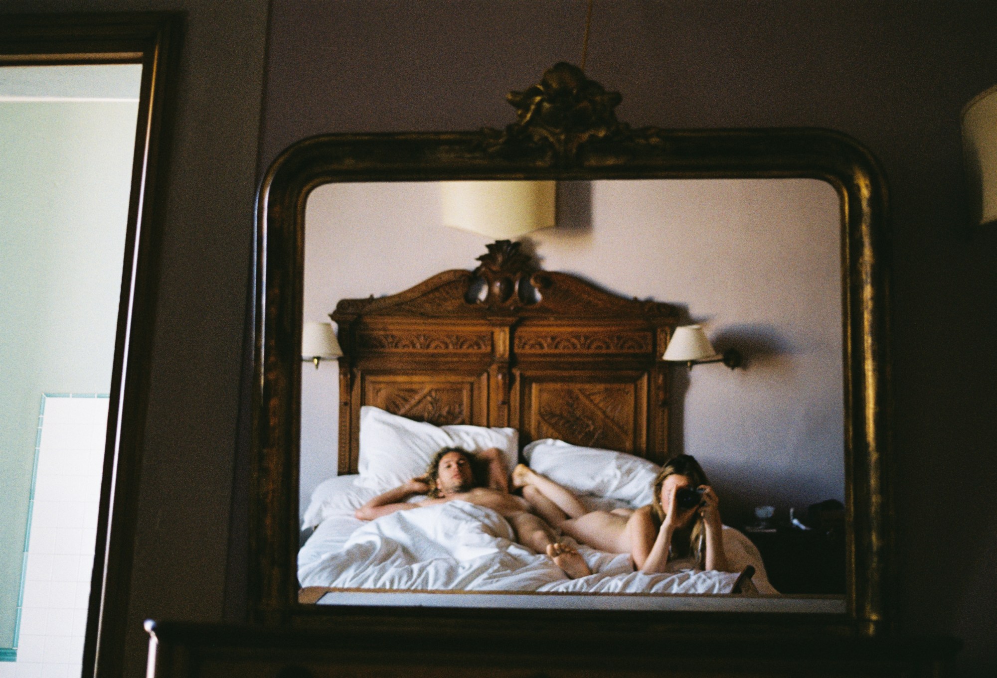 a nude couple lounge in an ornate wooden bed, one of them takes a photograph of the scene in a mirror