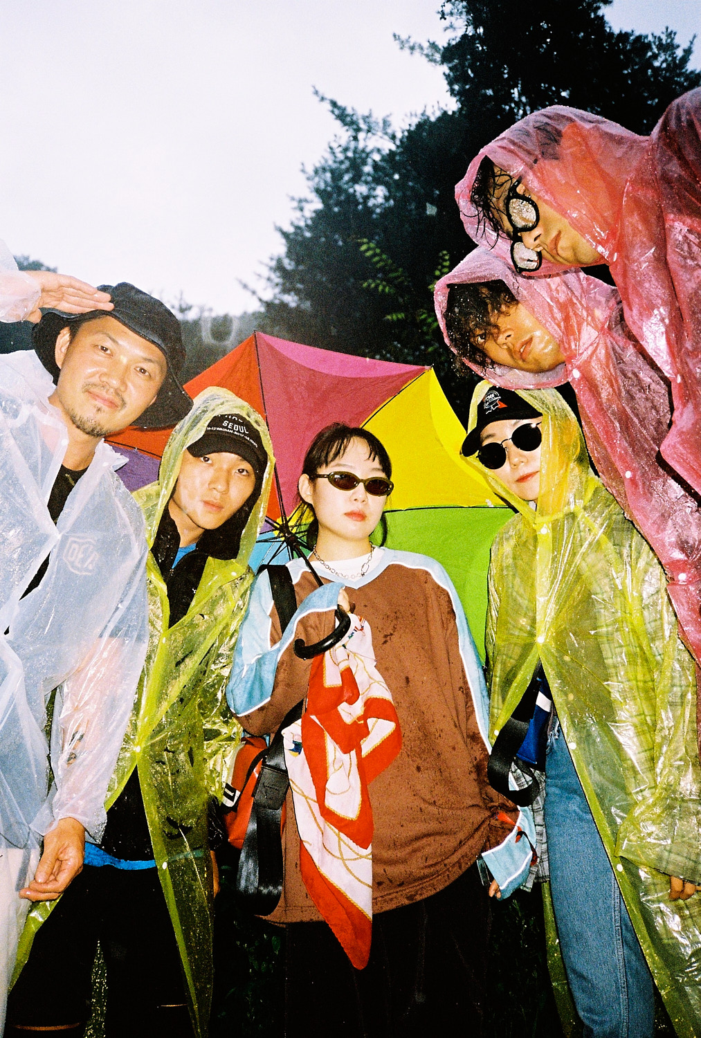 a korean band wearing colourful rain ponchos stand in the rain around the camera