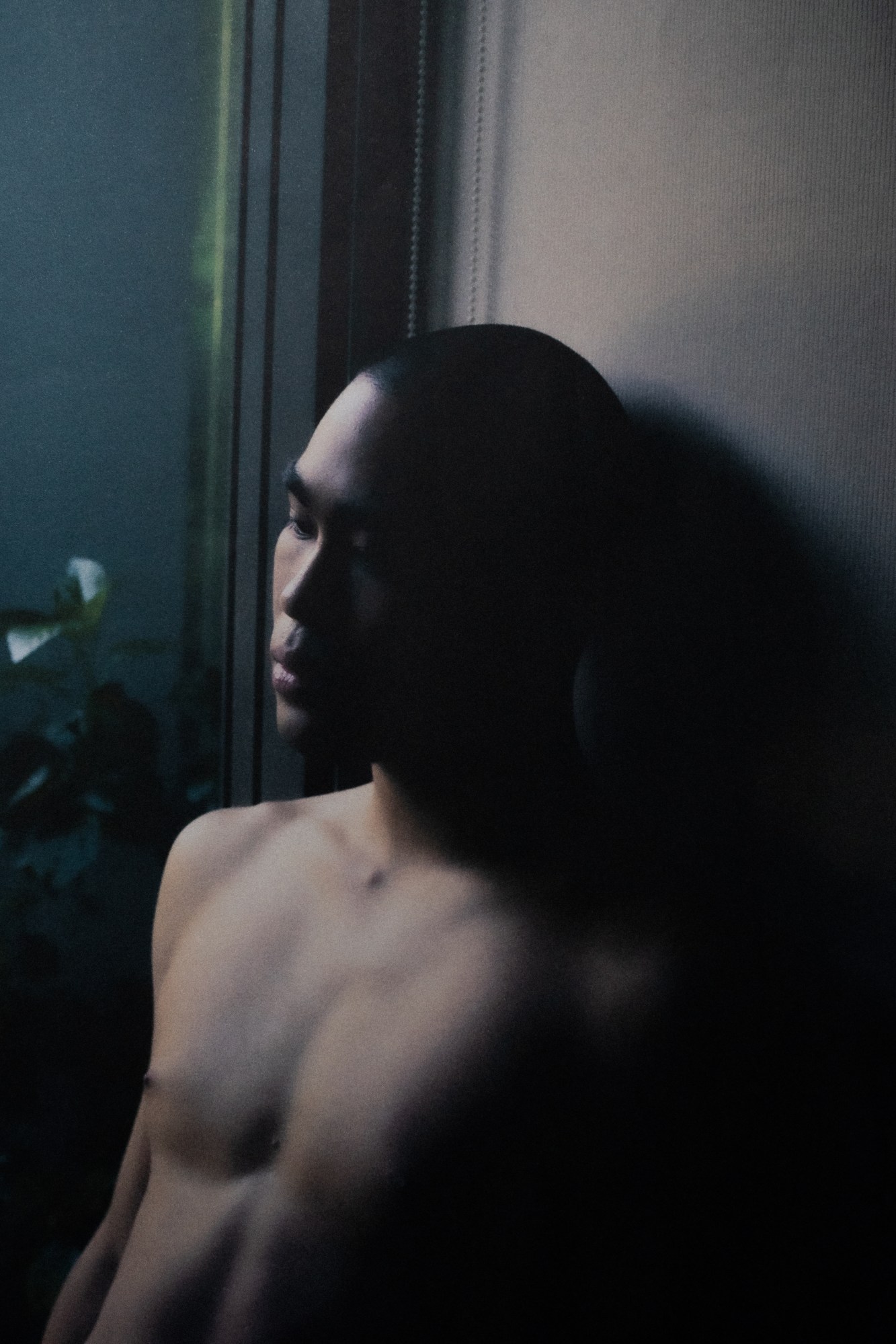 a topless man rests against a wall with green plants in the background