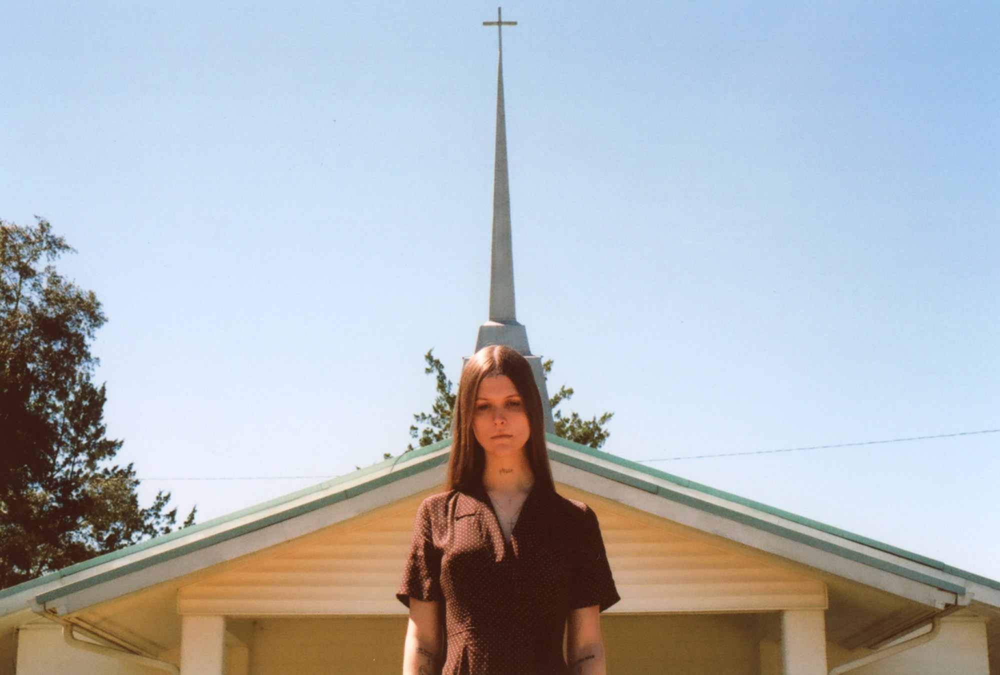 ethel cain in a brown collared dress, standing in front of a chapel in the sun