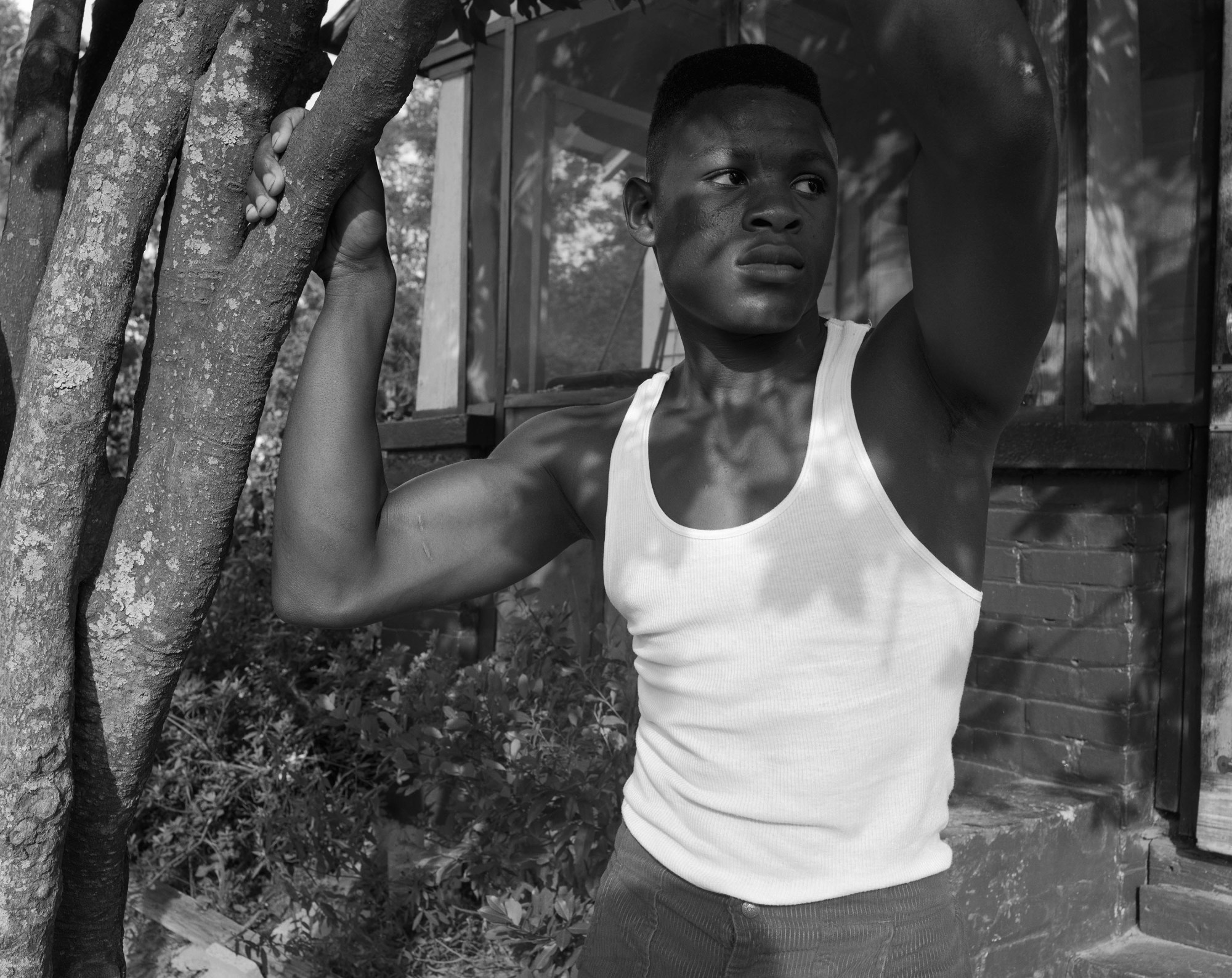 black-and-white photo by Baldwin Lee of a boy in a white vest holding onto a tree outside a home.