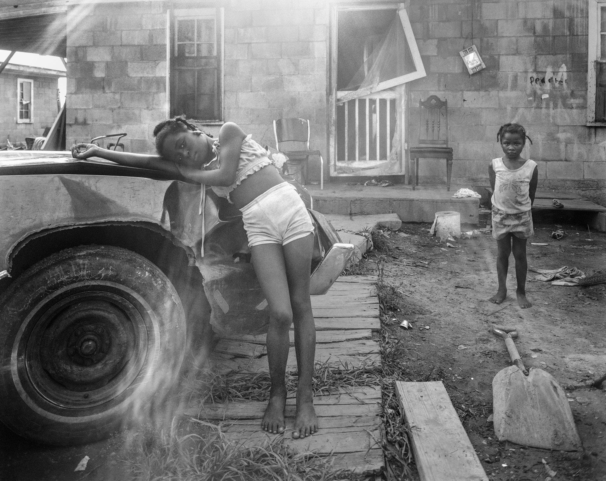 black-and-white photo by Baldwin Lee of a little girl leaning on a tractor while another stands in the background outside a deserted and destroyed home.