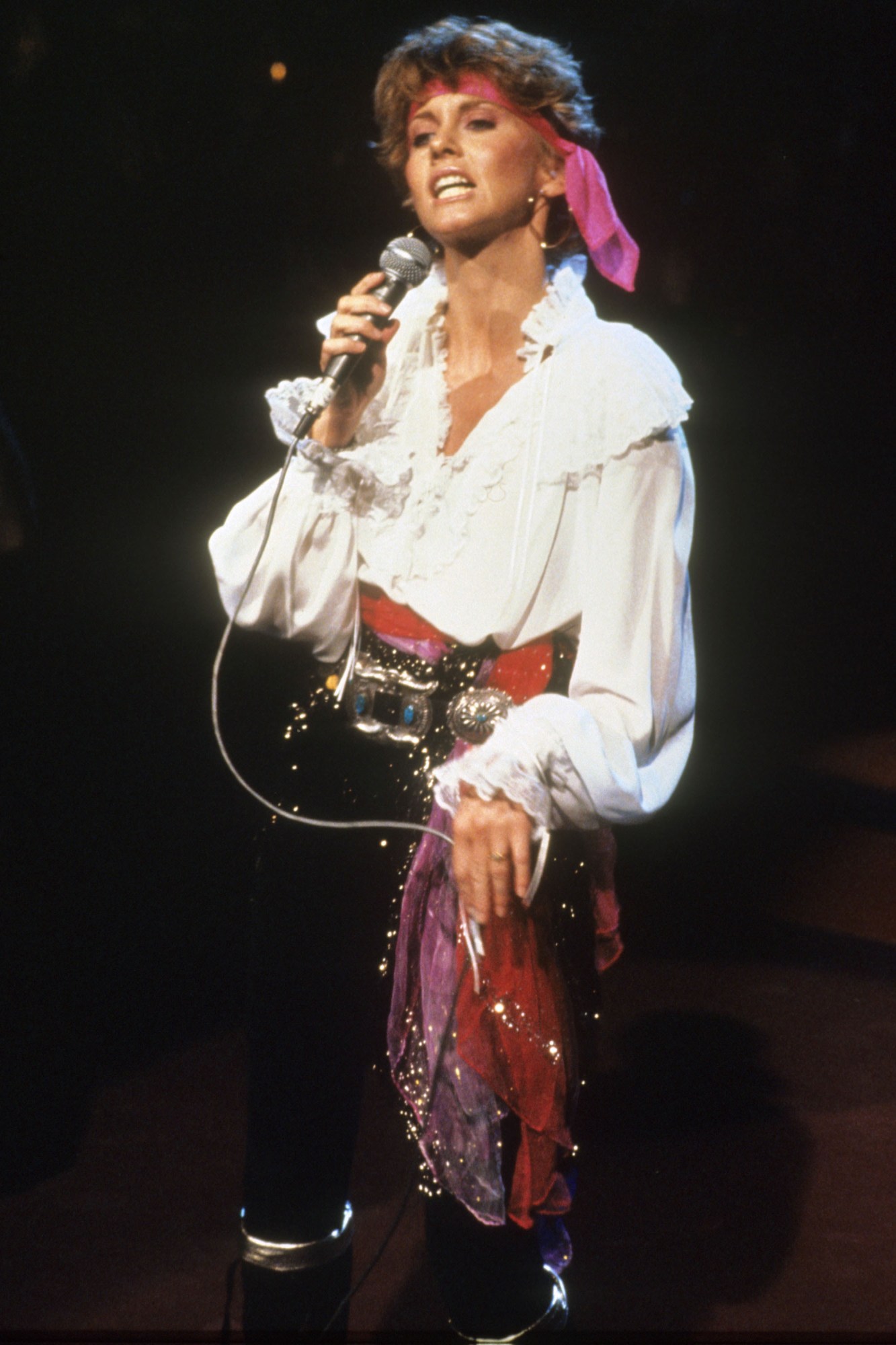 Olivia in a pirate costume, with pink scarves tied around her head and waist, singing on stage