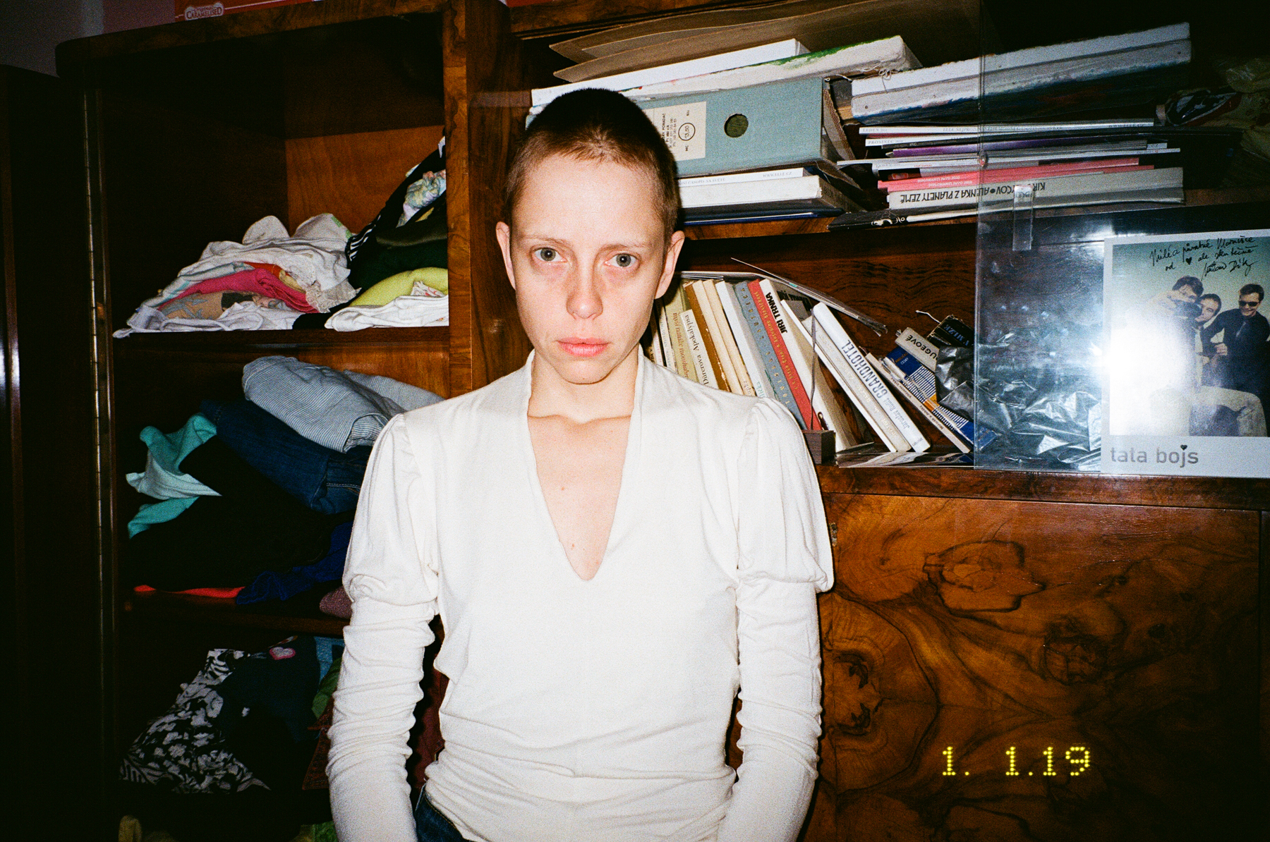 Photograph by Marie Tomanova of herself wearing her old clothes in her old room with books, clothes and records in the cupboards behind.