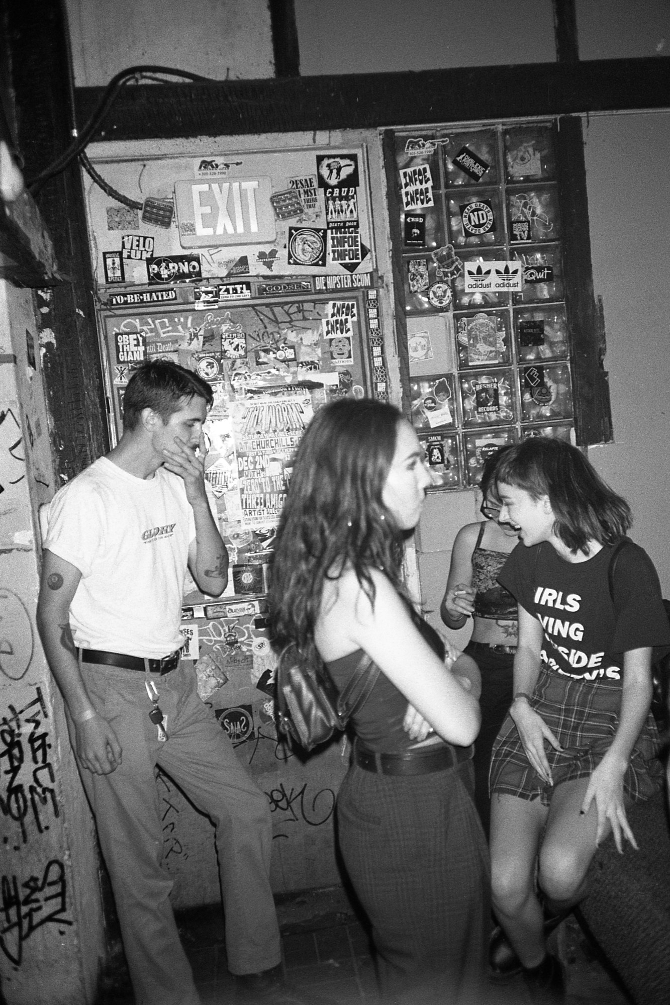 Black-and-white photograph of club goers hanging around a wall covered in stickers, posters and graffiti