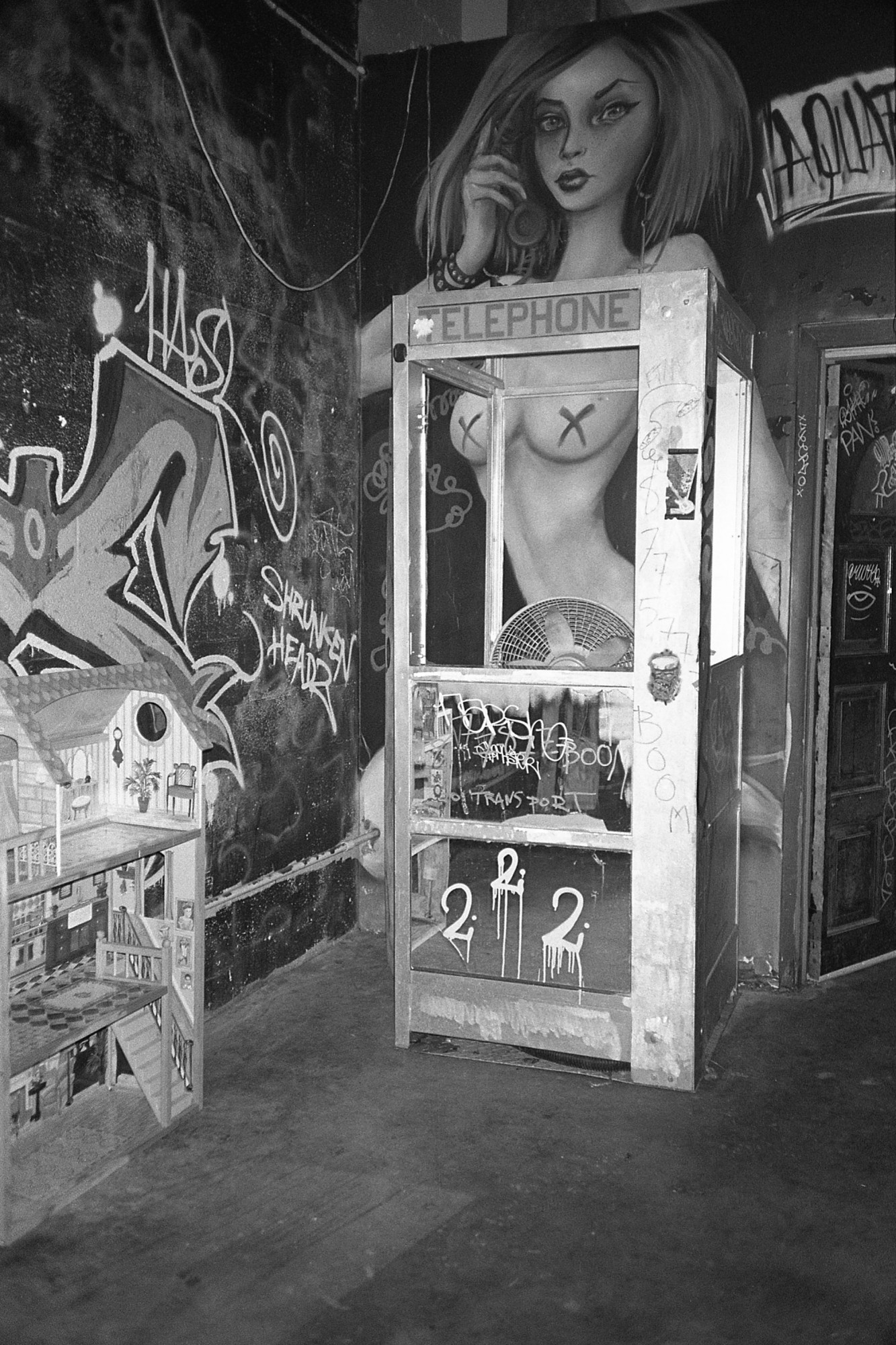 Black-and-white photograph of a club with paintings of naked women and graffiti on the wall, a telephone booth sits against one wall and a doll house against the other.