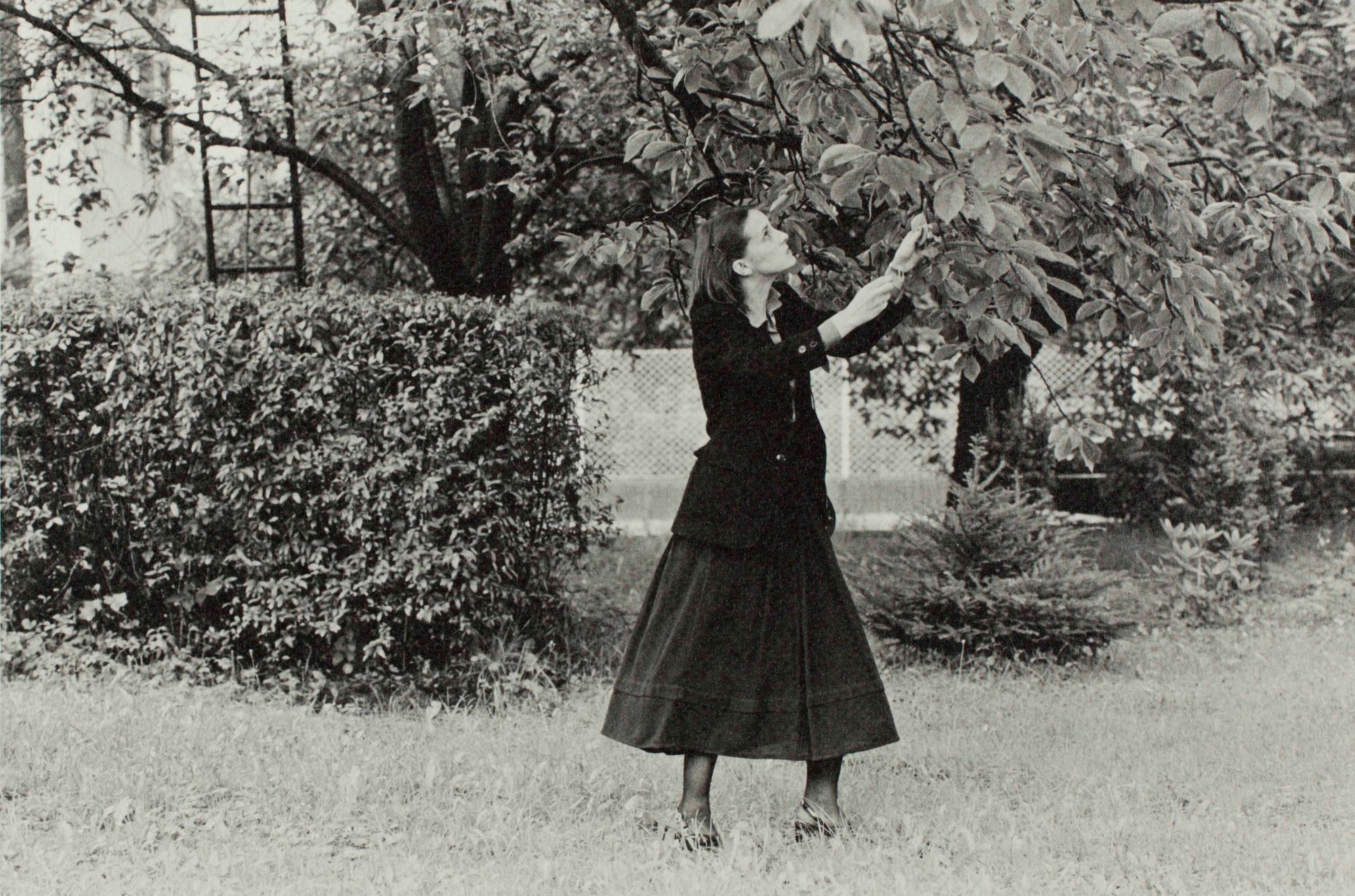 black and white image of a woman in a black skirt and black jacket in a garden touching a tree