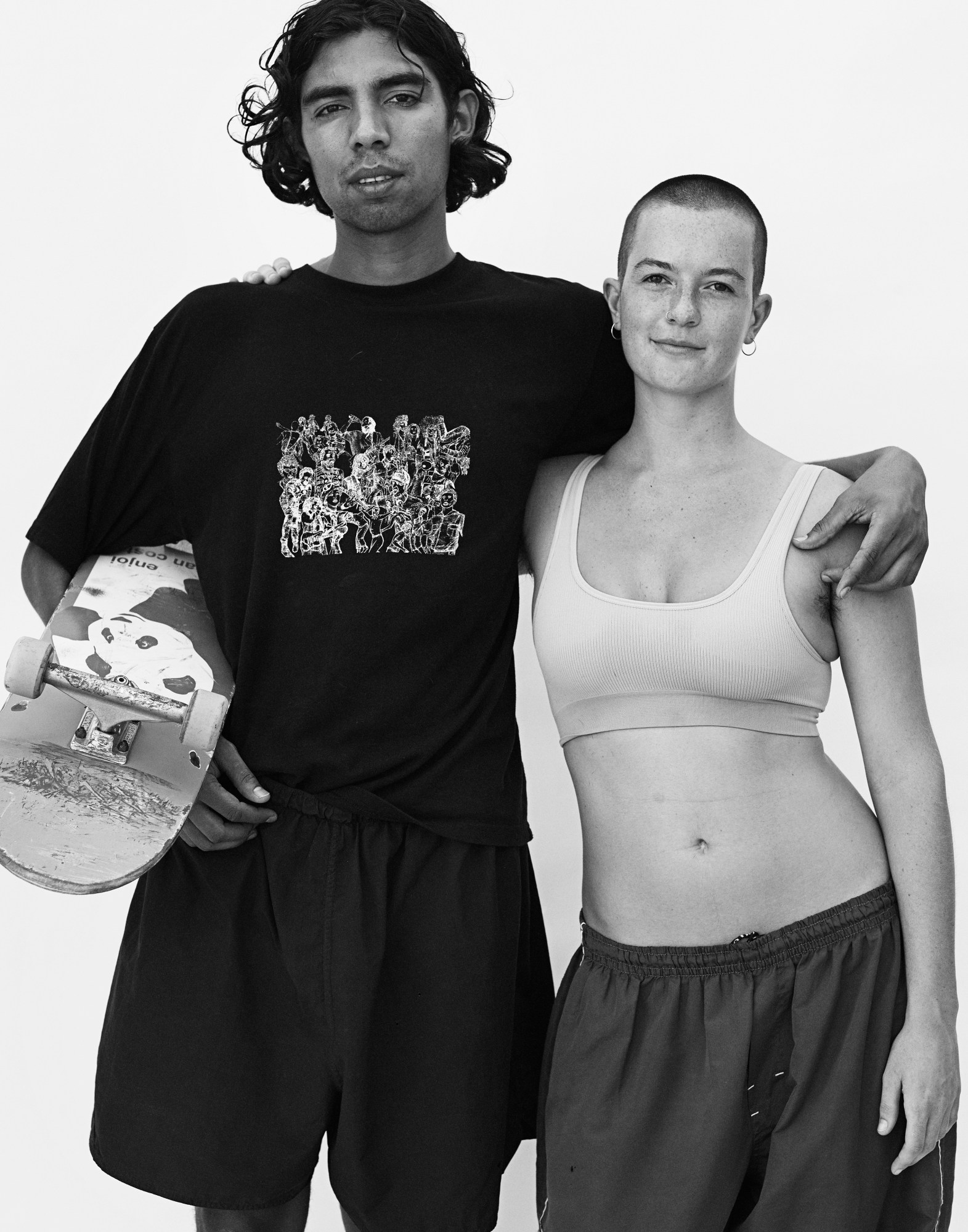 a couple stand with their arms around each other, the model on the left holding a skateboard