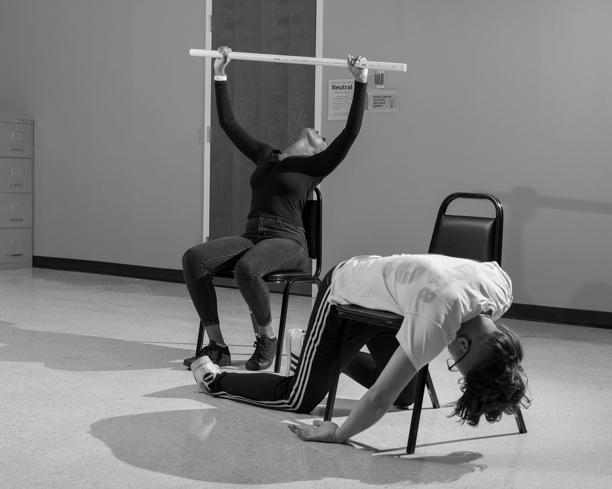 two people in a church basement on chairs, one lying back holding a pole above their head, the other slouched over one