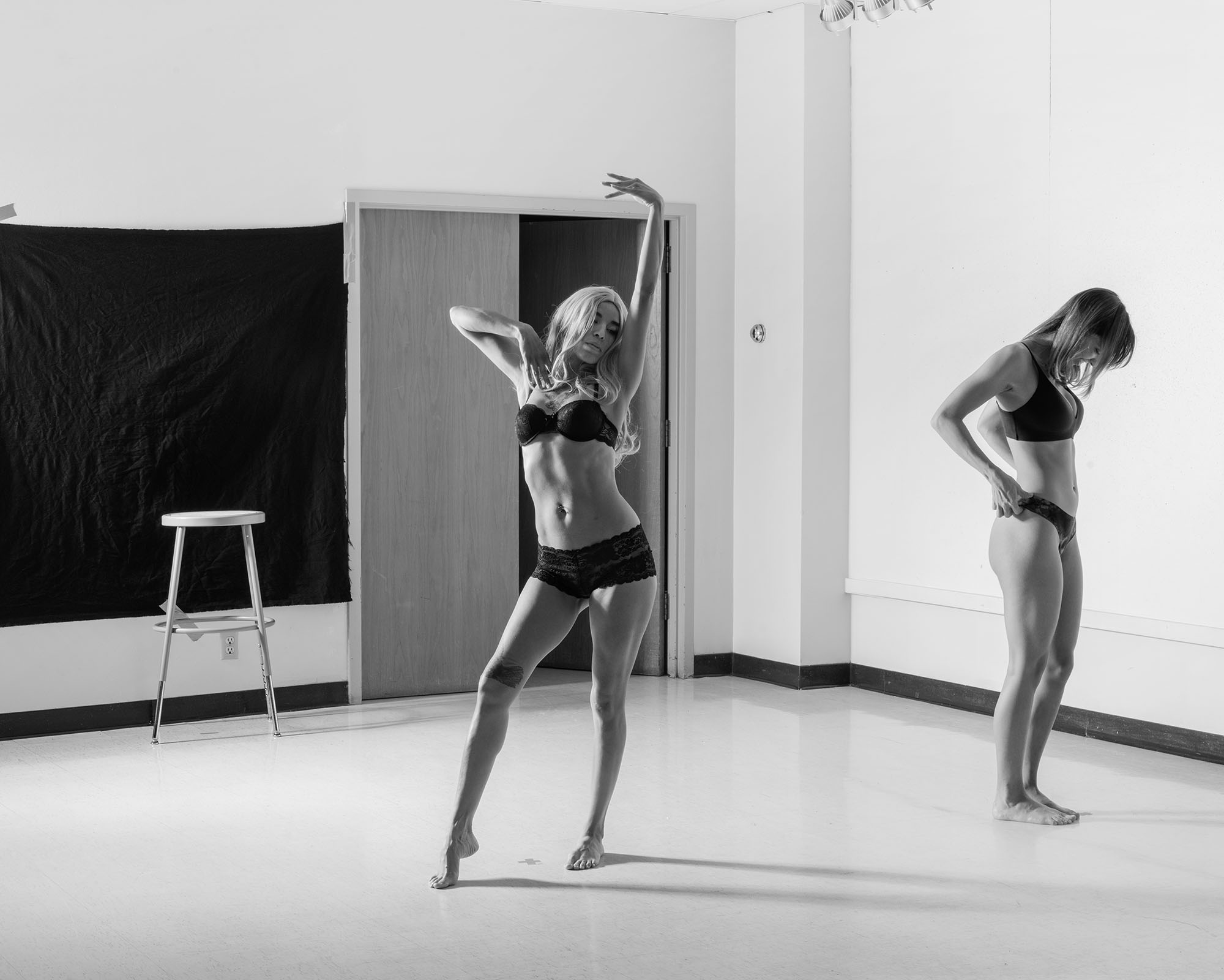 two people in a church basement in their underwear, one posing with her toes pointed, the other looking to the ground