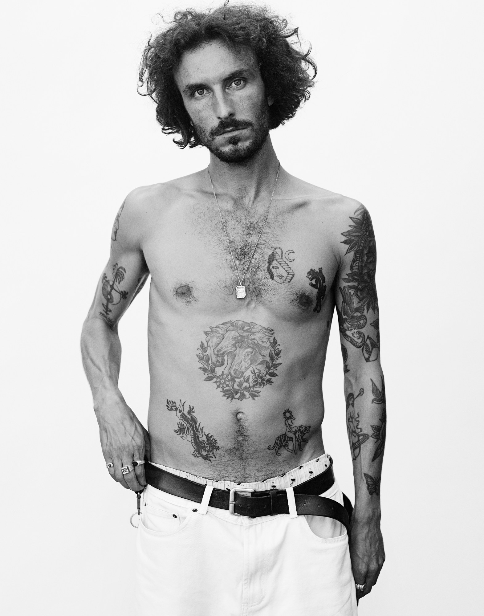 a portrait of a shirtless model wearing white trousers with a number of tattoos on his torso