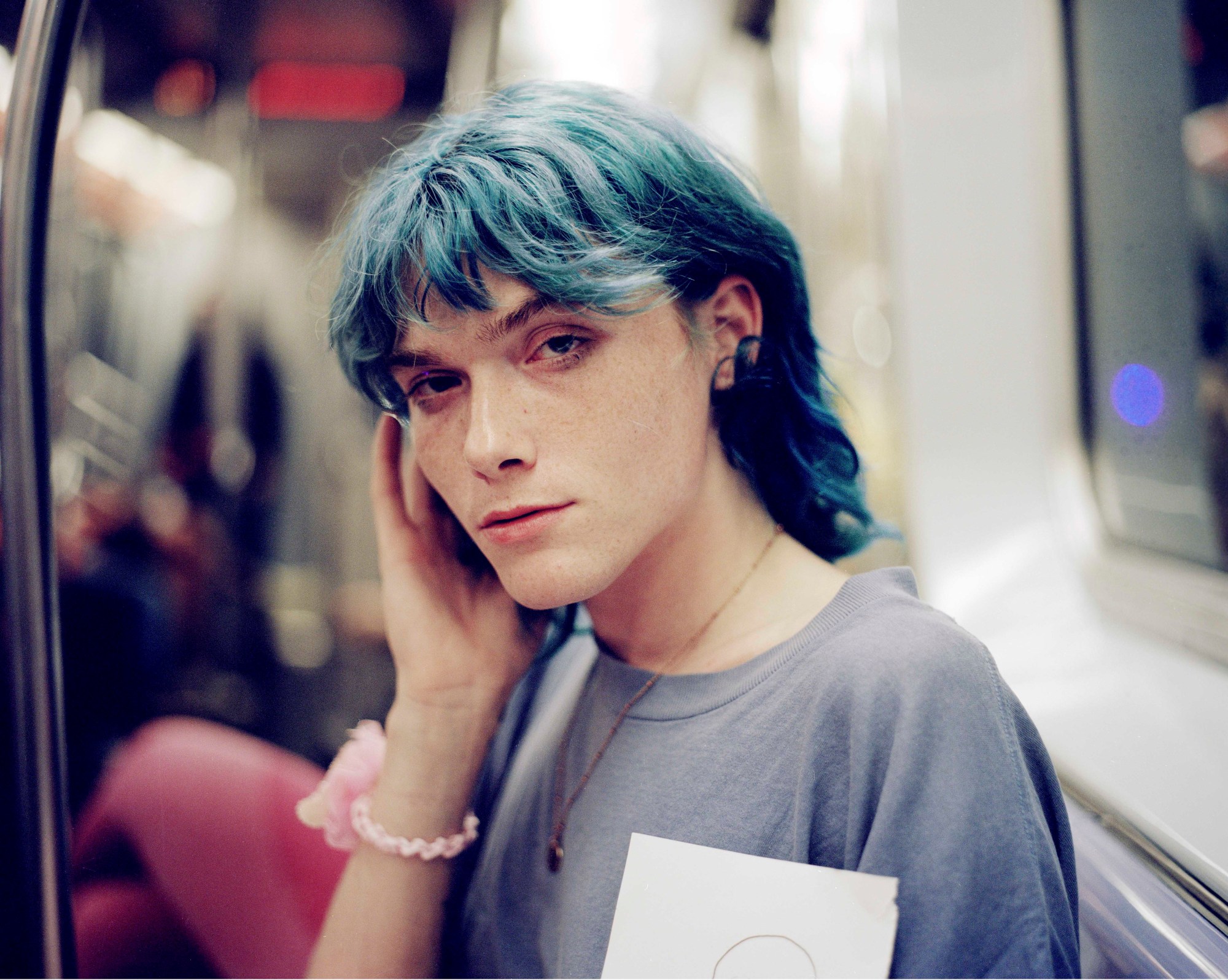 a person with a blue mullet sitting on an underground train cups their face and looks to camera