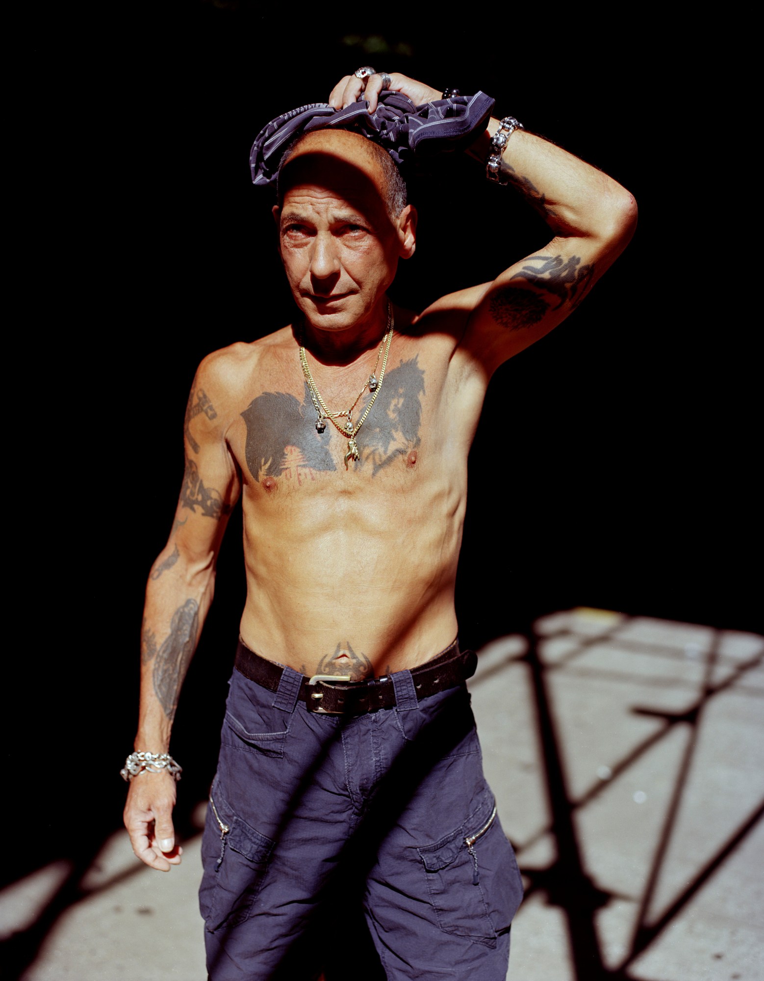 an older tattooed man stands topless in the sunlight mopping his head with a bandana