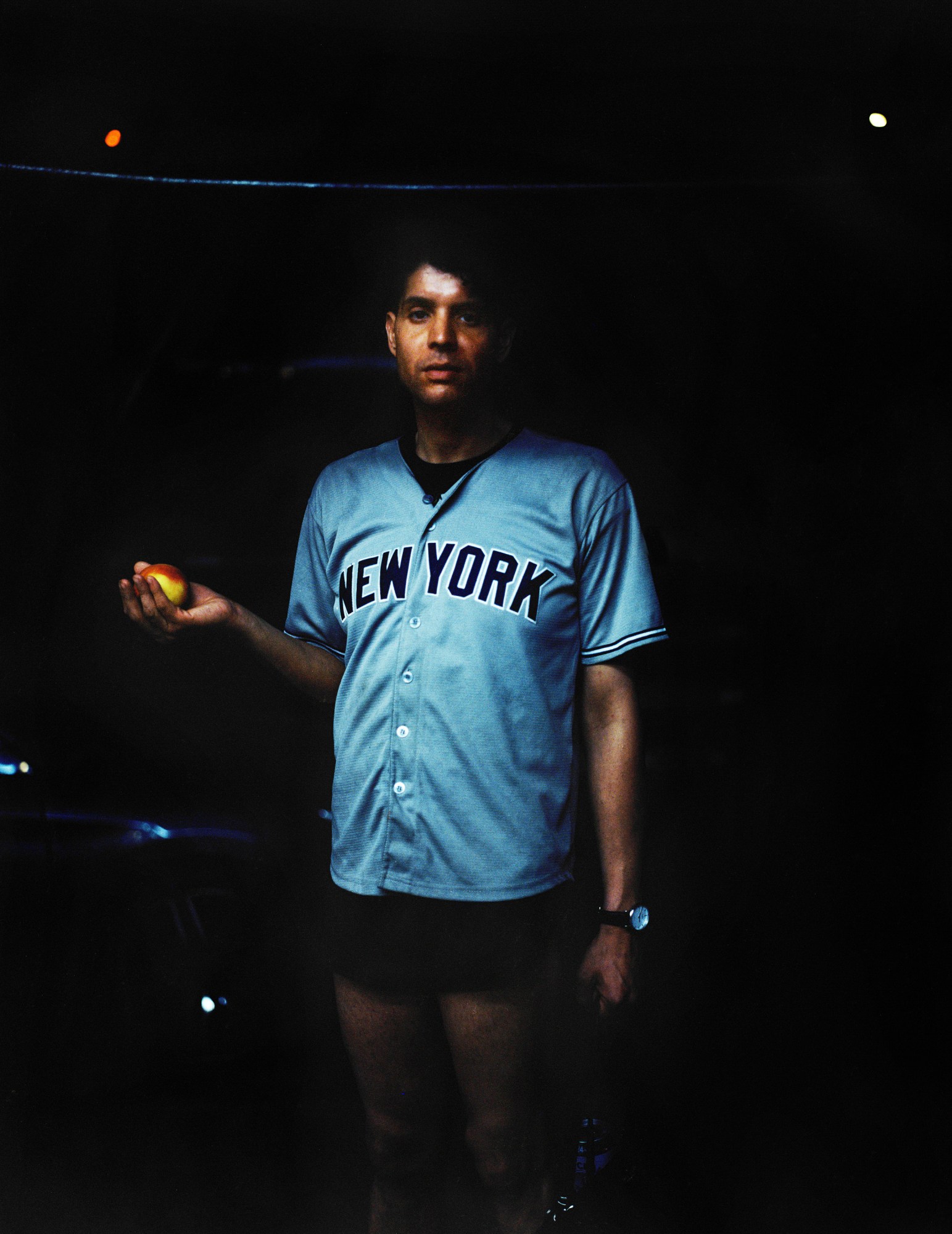 a man holding an apple stands in the dark wearing a baby blue New York jersey and short shorts