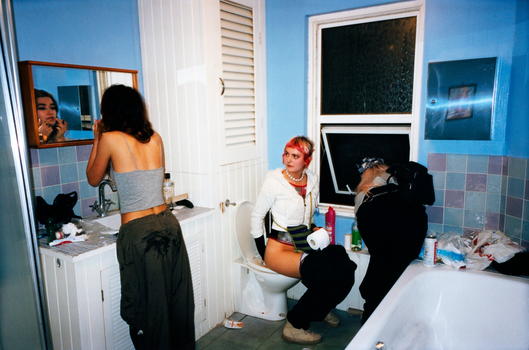 Photo by Evie Shandilya of three friends in a bathroom, one on the toilet, another applying makeup and one looking out the window in the book 'I Just Want to Party'