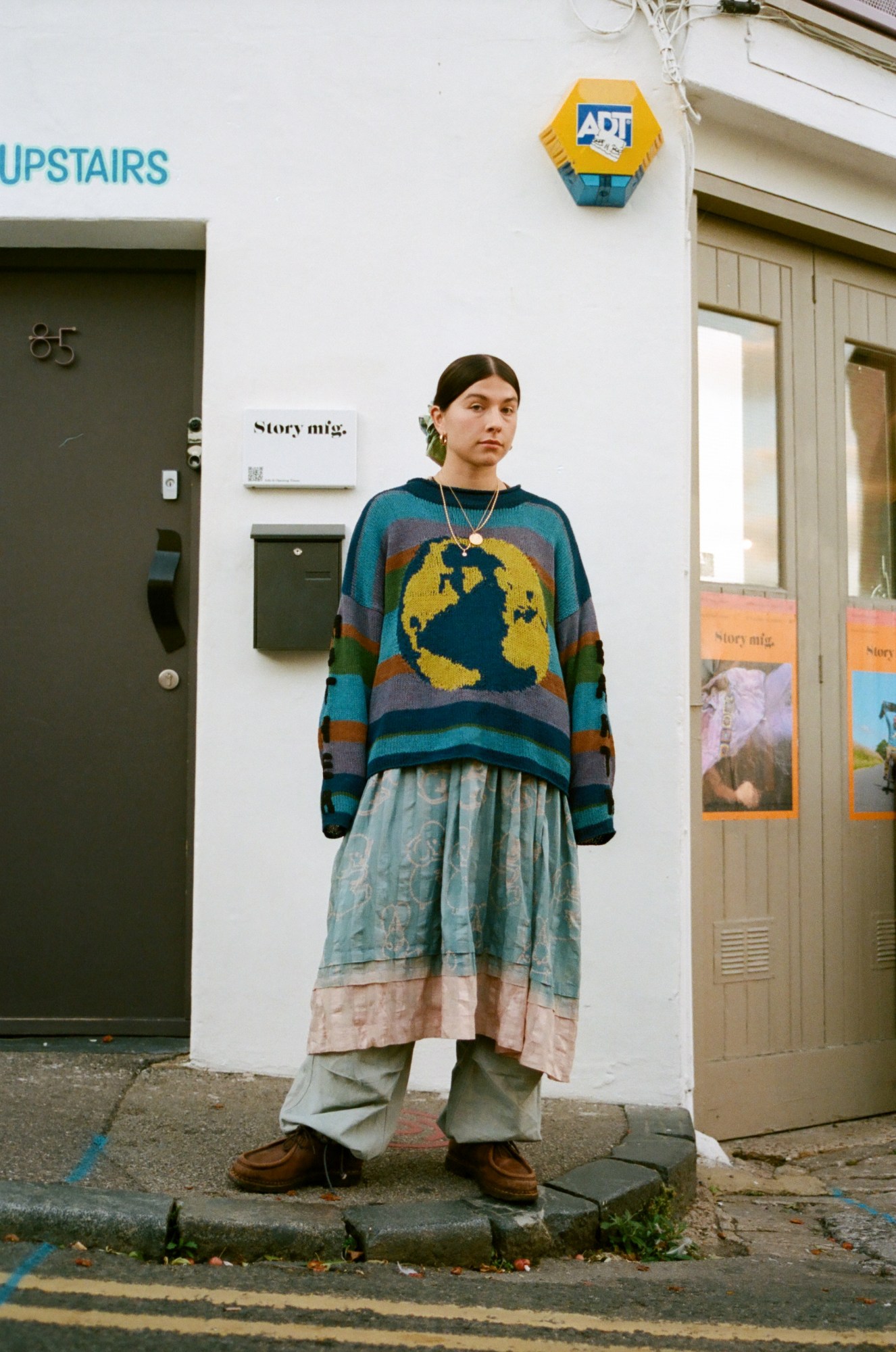 a brunette woman stands in the street wearing a knitted globe jumper over a dress and trousers