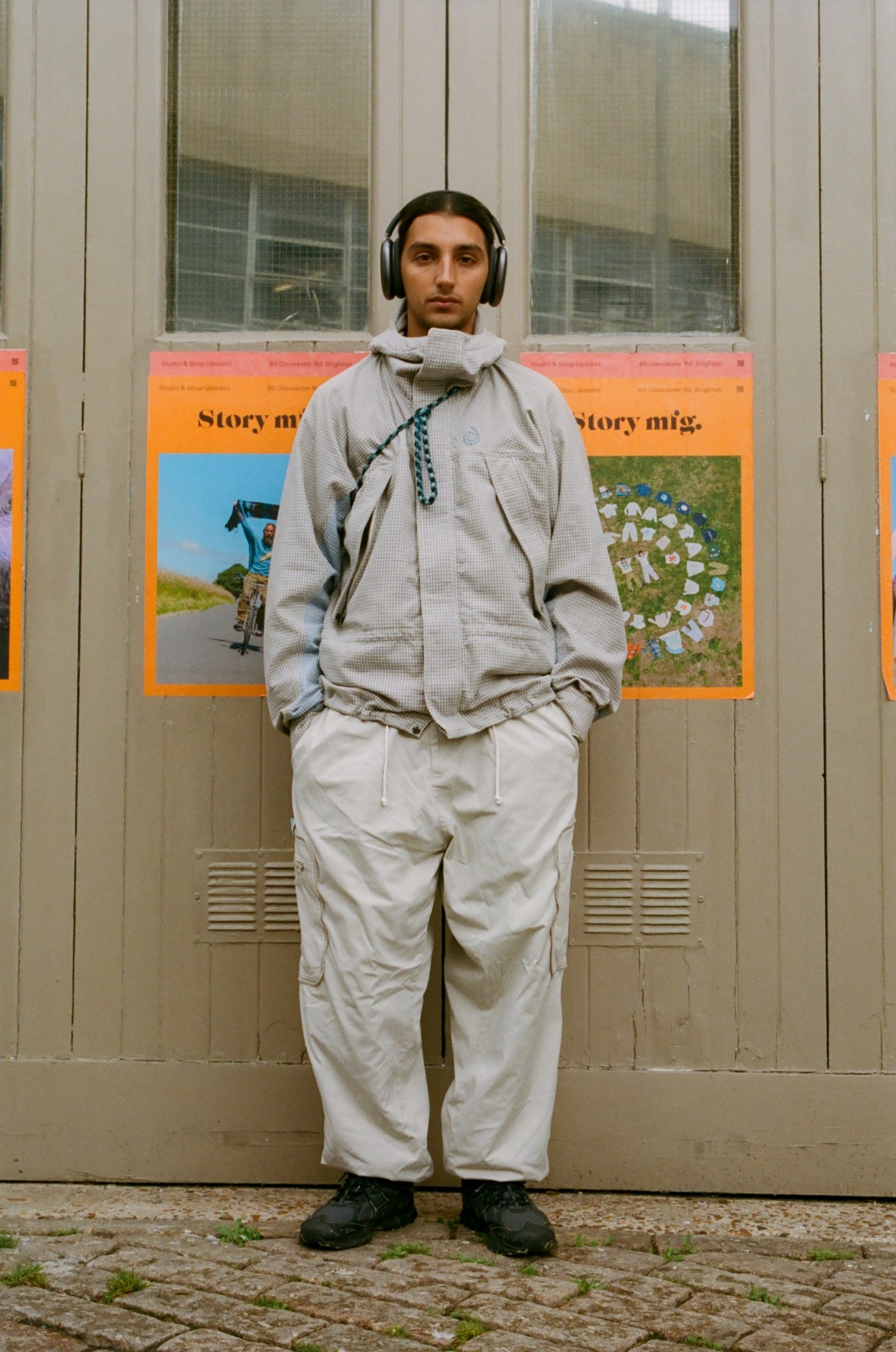 a man wearing headphones with a beige jacket and combat trouser combo stands in the street