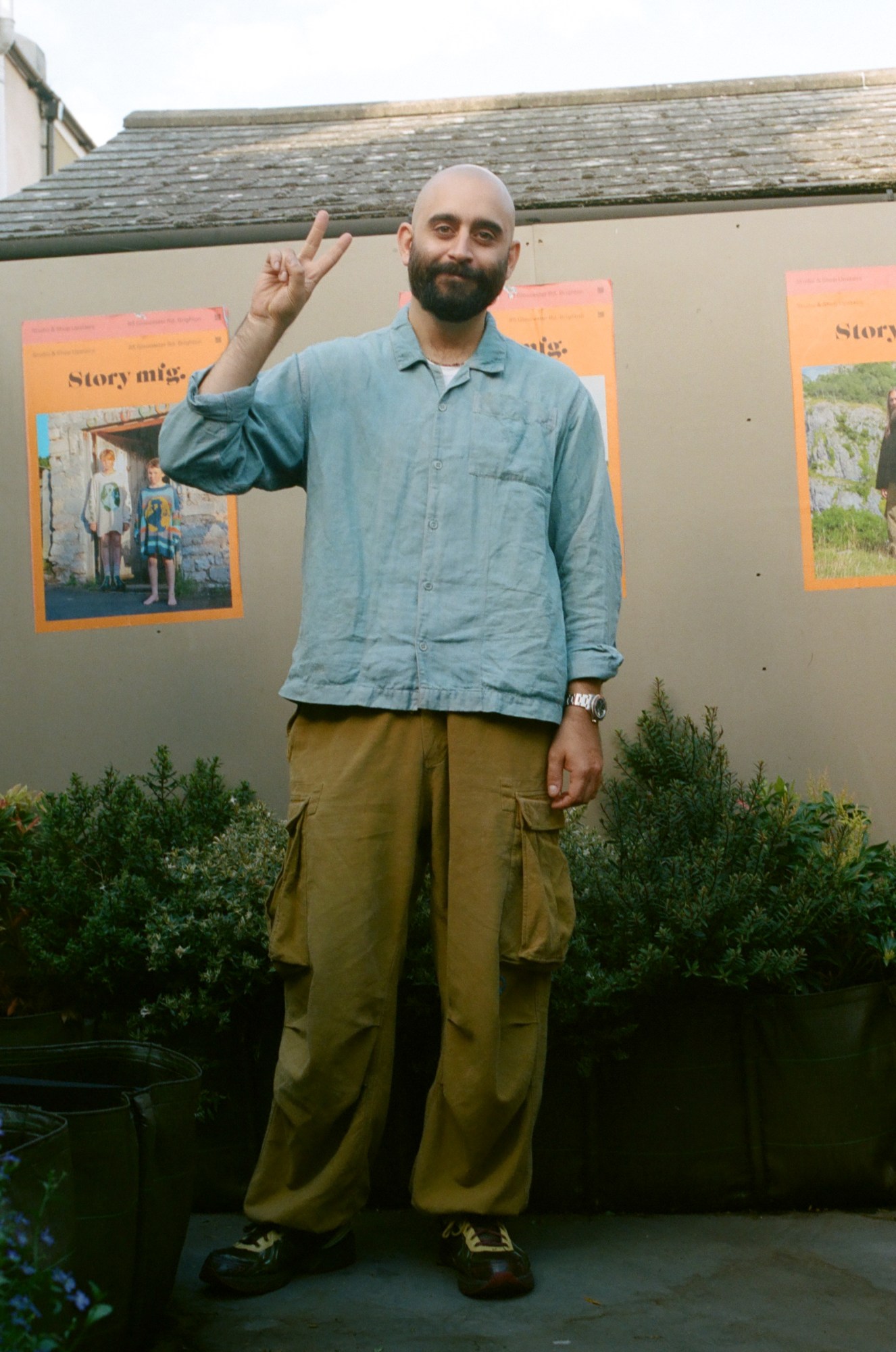 a man in combat trousers and a blue shirt stands surrounded by plants giving the peace sign