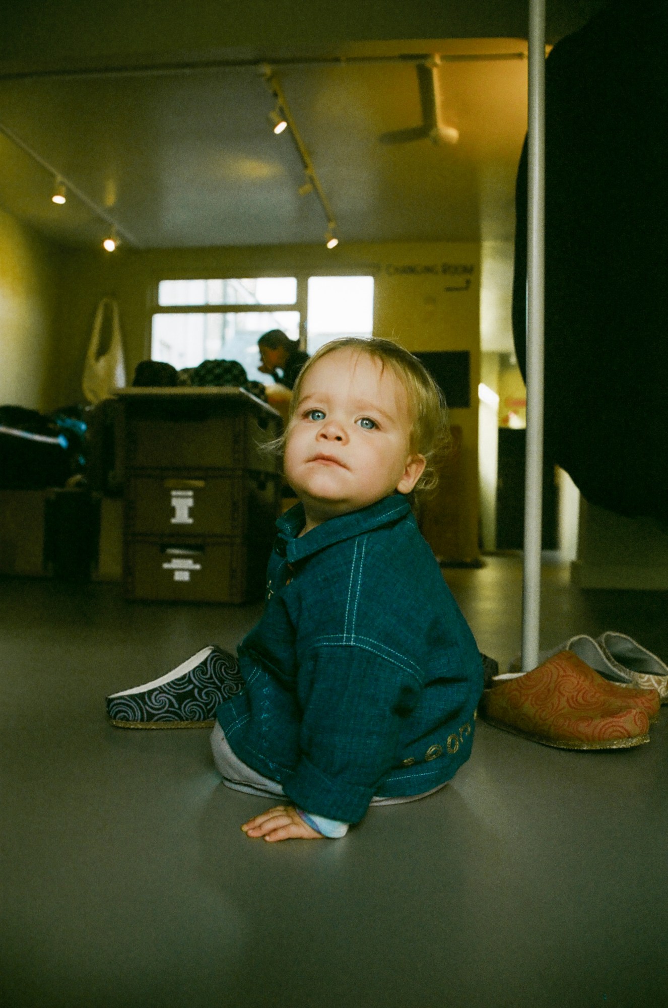 a baby in a denim shirt sits on the shop floor surrounded by clogs