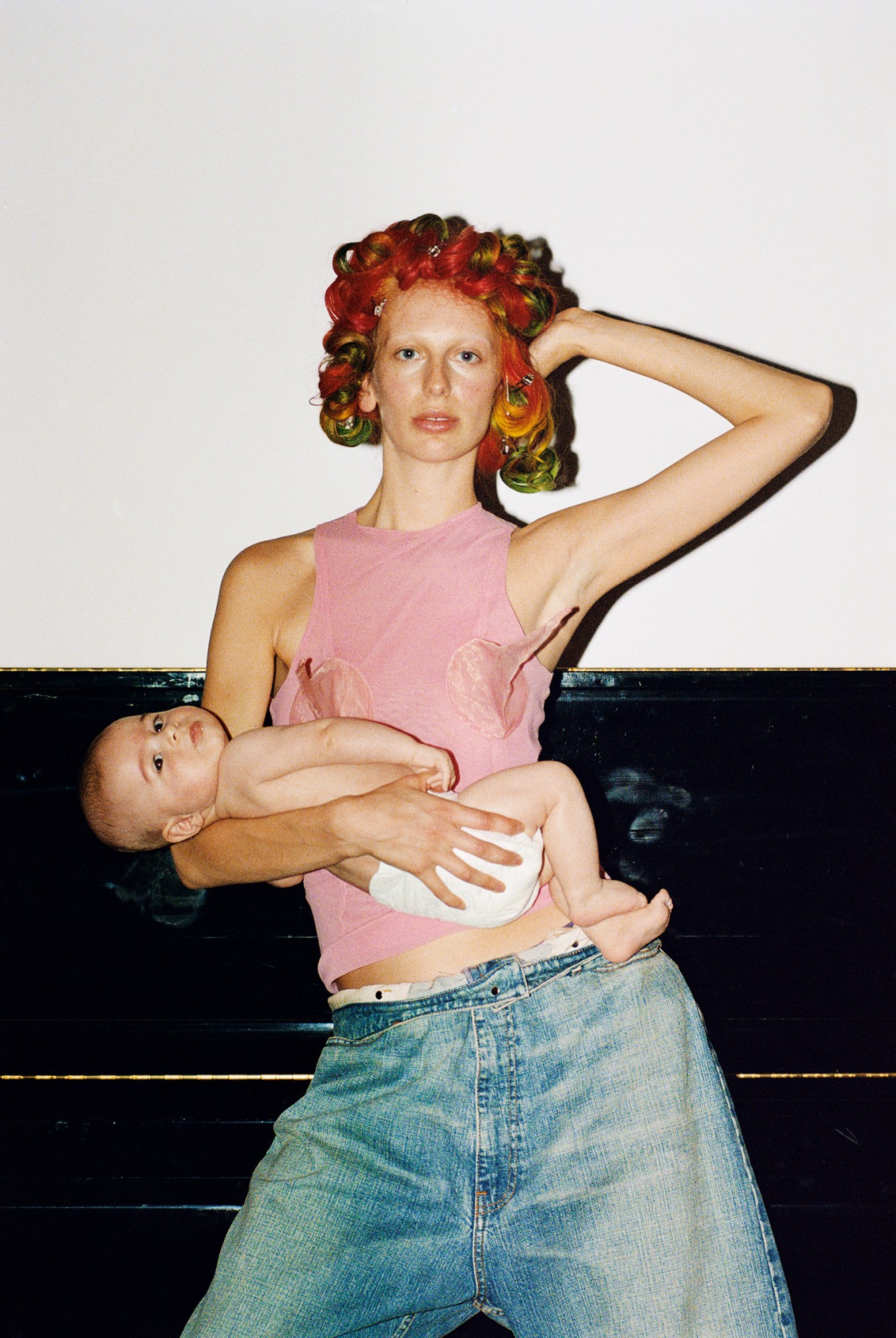 A woman holding a baby while wearing a pink tank top and jeans.