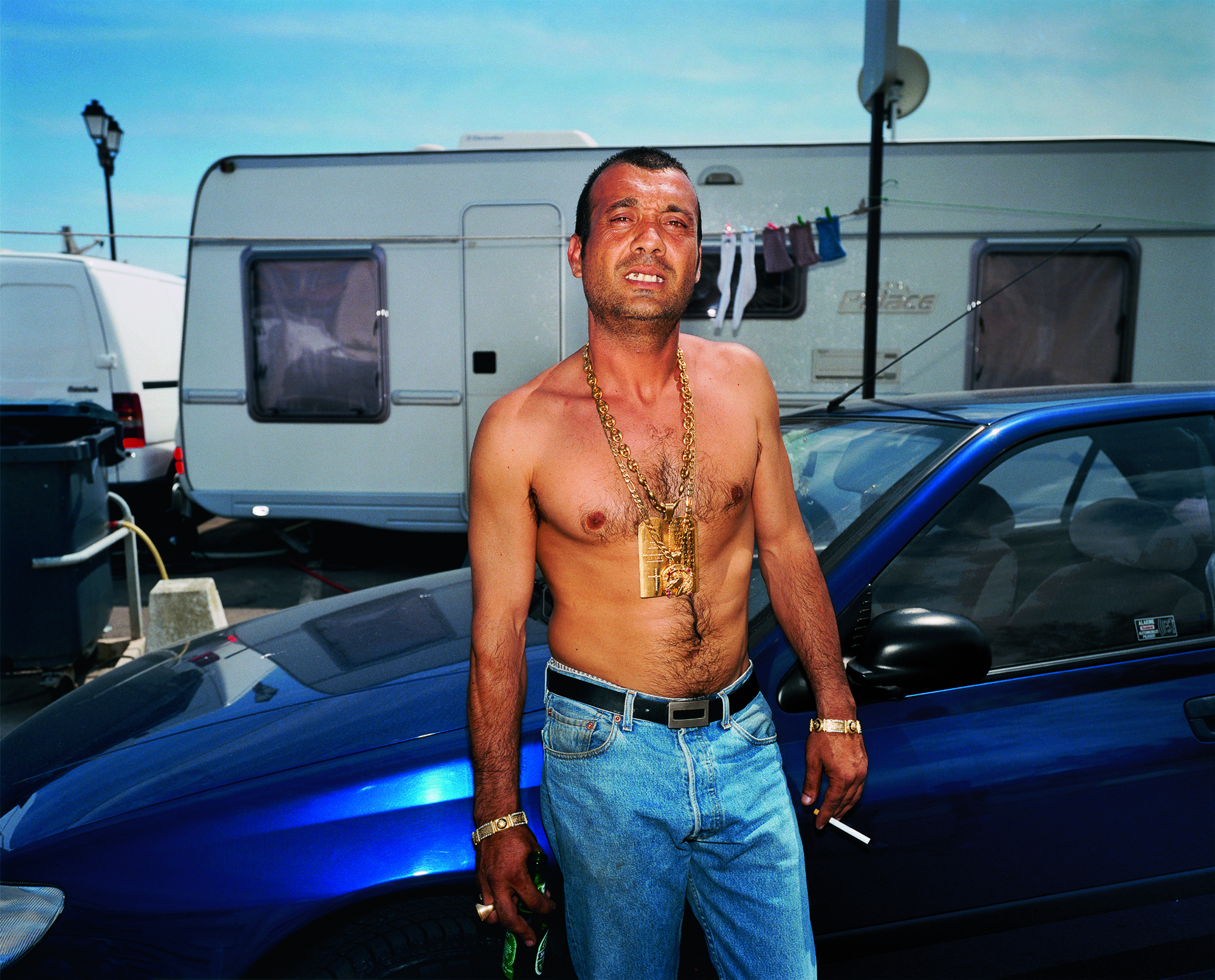 a topless man in blue jeans with gold jewellery smoking in front of a blue car