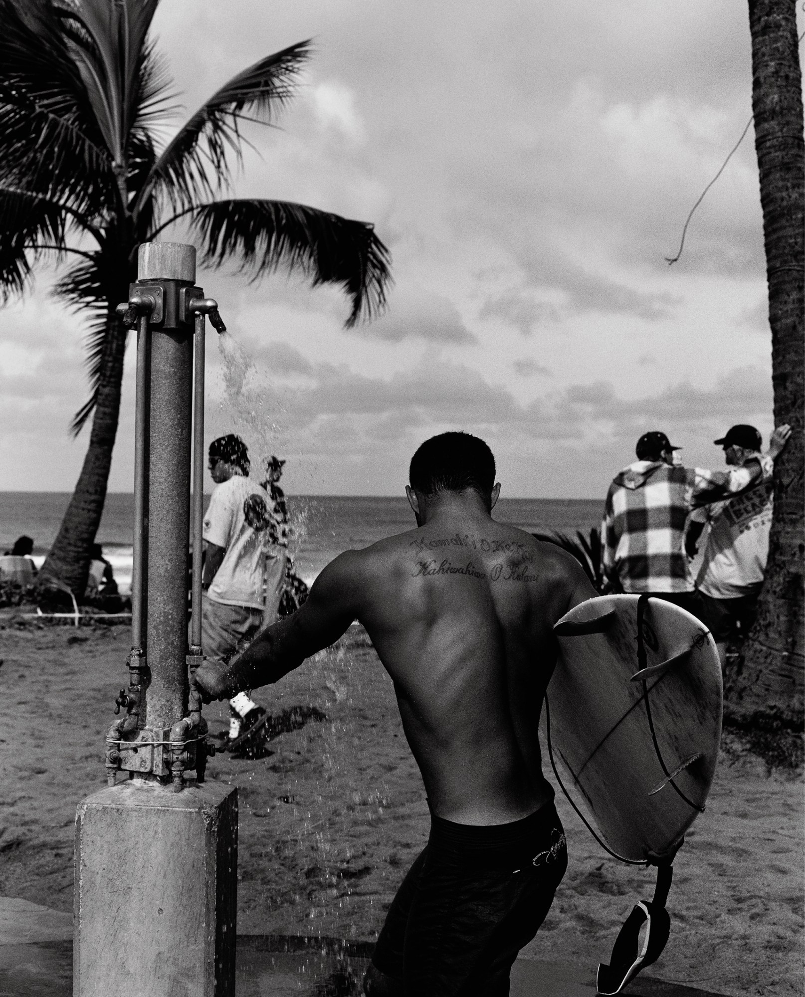 sepia toned image of a topless man showering on the beach while holding a surfboard