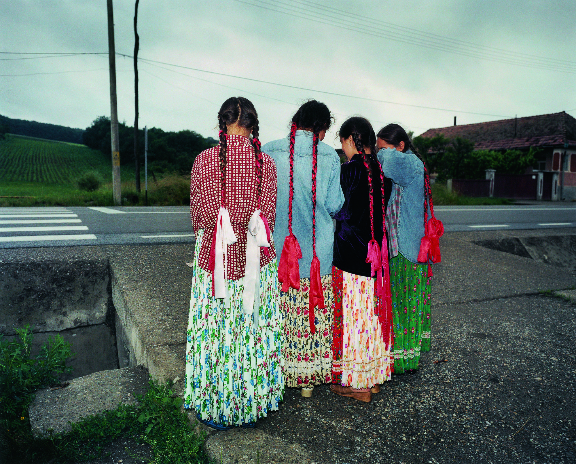 young girls stand facing away from the camera on the side of the road, their hair plaited with red ribbons