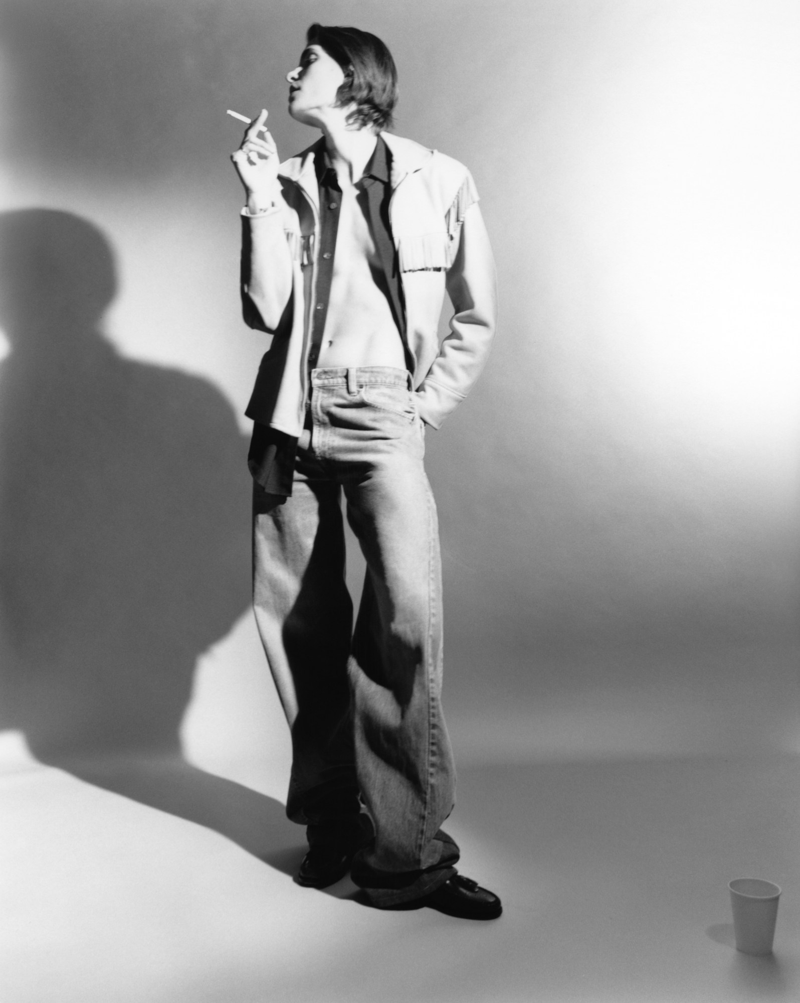 a model wears an open shirt and jacket and a pai of denim jeans, smoking a cigarette in a photography studio