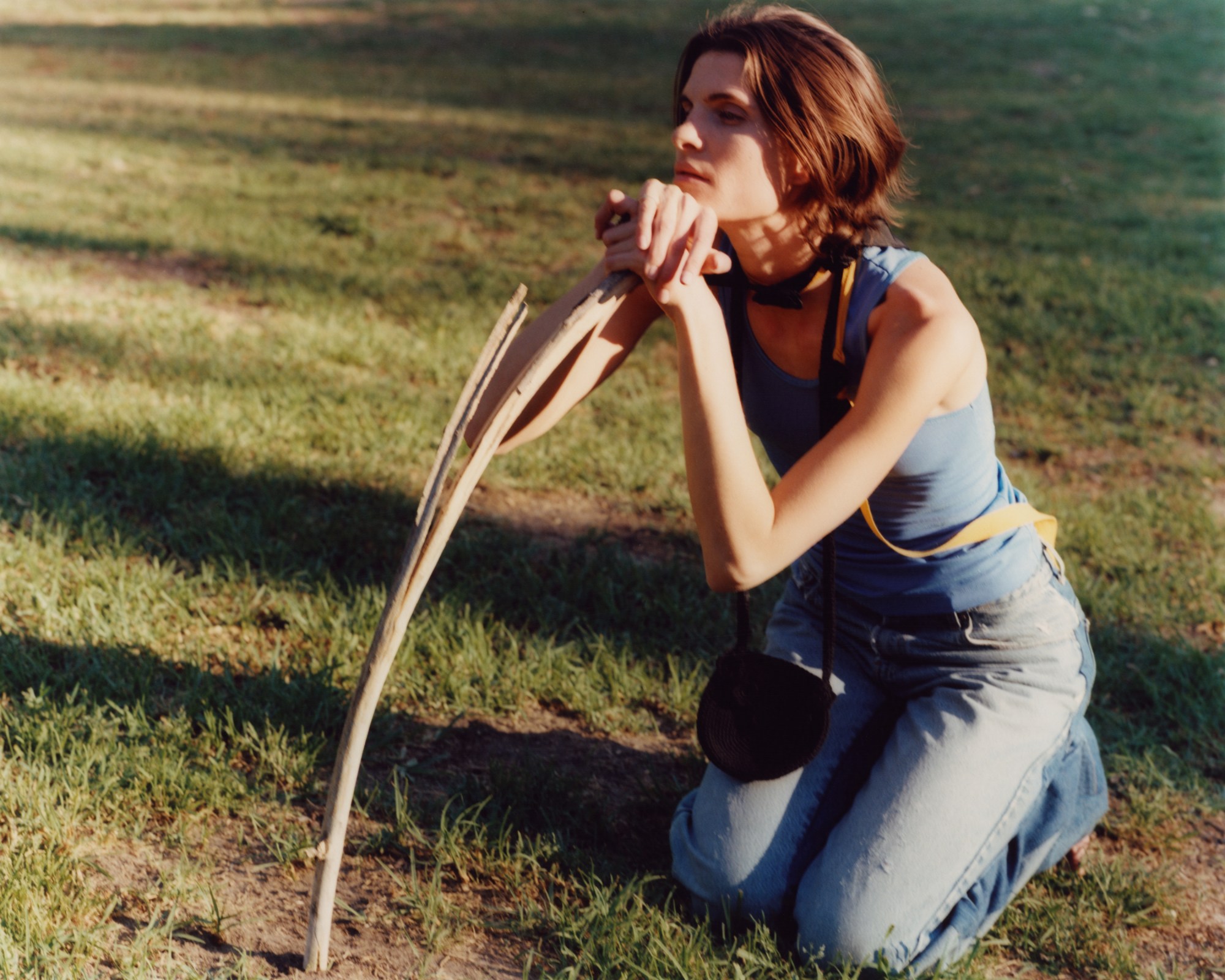 a model wearing jeans and a tank top kneels in muddy grass outside, chin resting on a stick