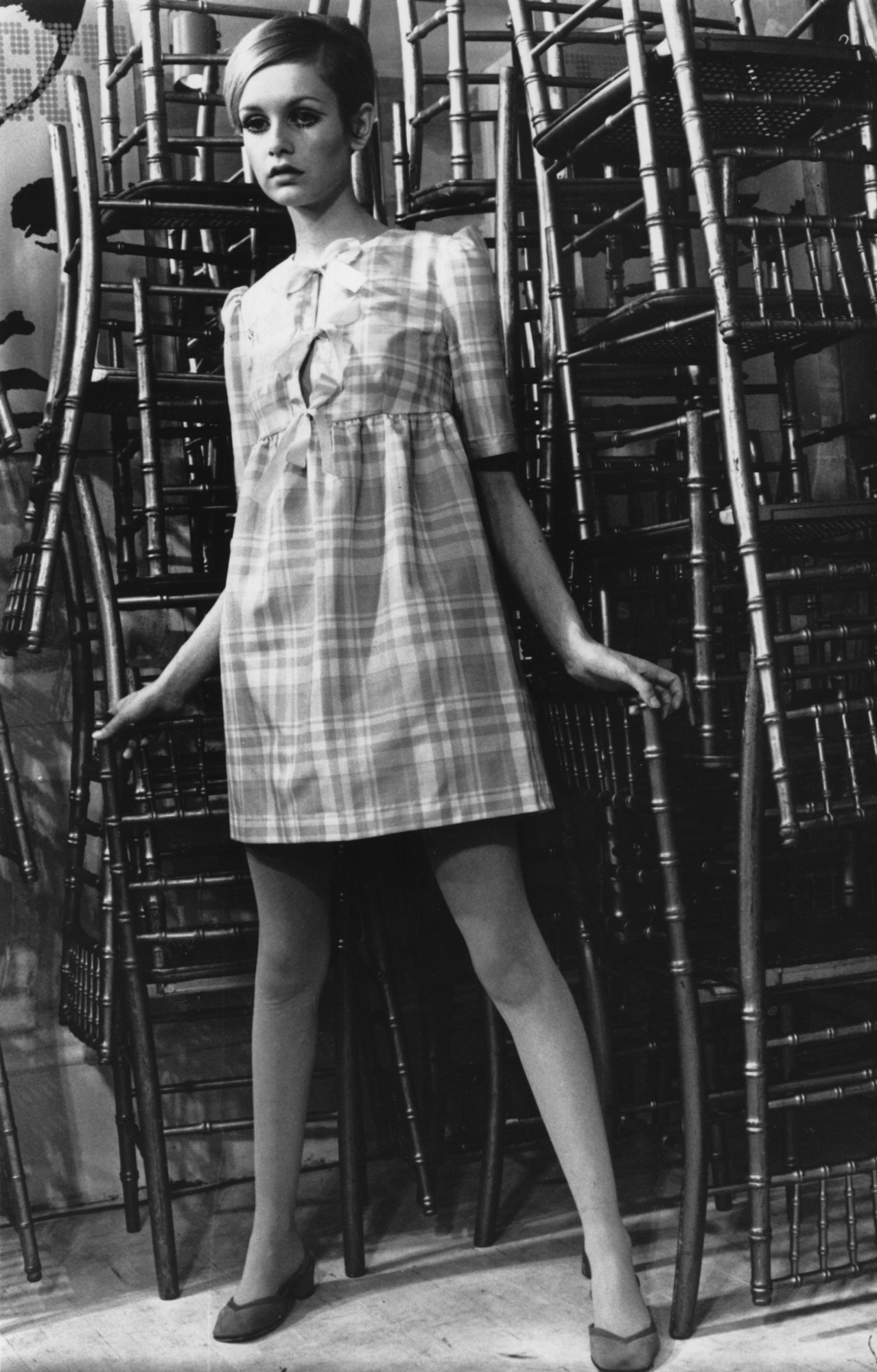 twiggy posing in a 60s swing dress she made in front of stacked wooden chairs 1967