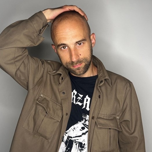 a man in khaki jacket and black slogan t-shirt against a white wall