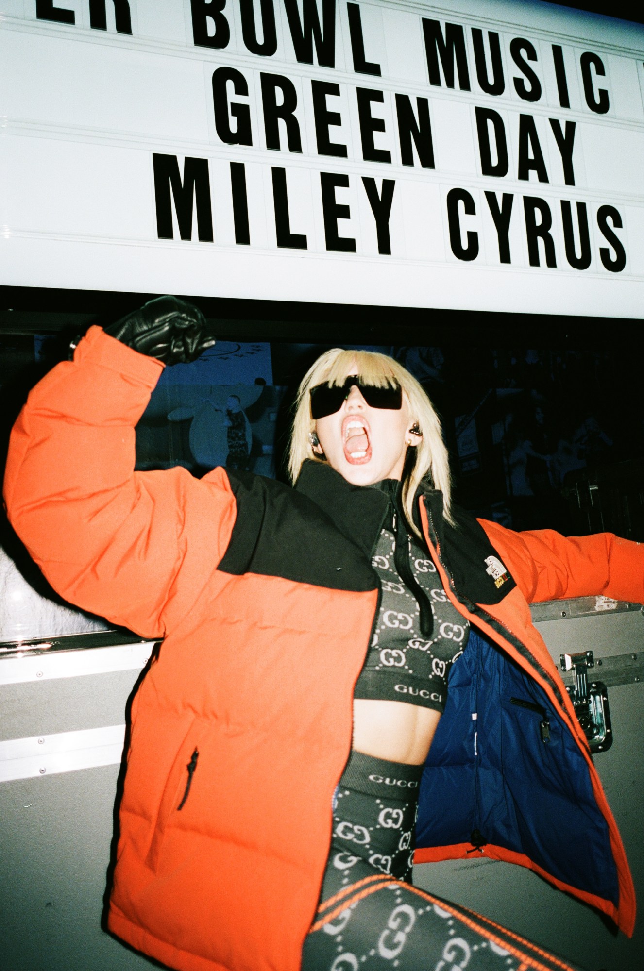 miley cyrus in an orange puffer jacket and dark glasses posing in front of a billboard with her name on it