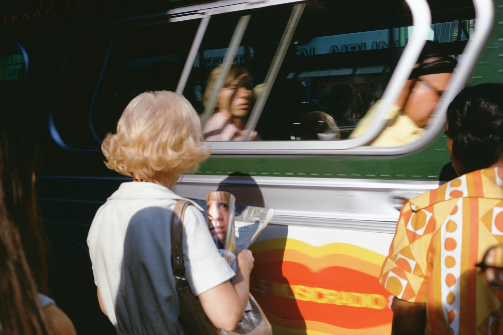 a blonde woman looking at a magazine in front of a new york city bus in the 70s