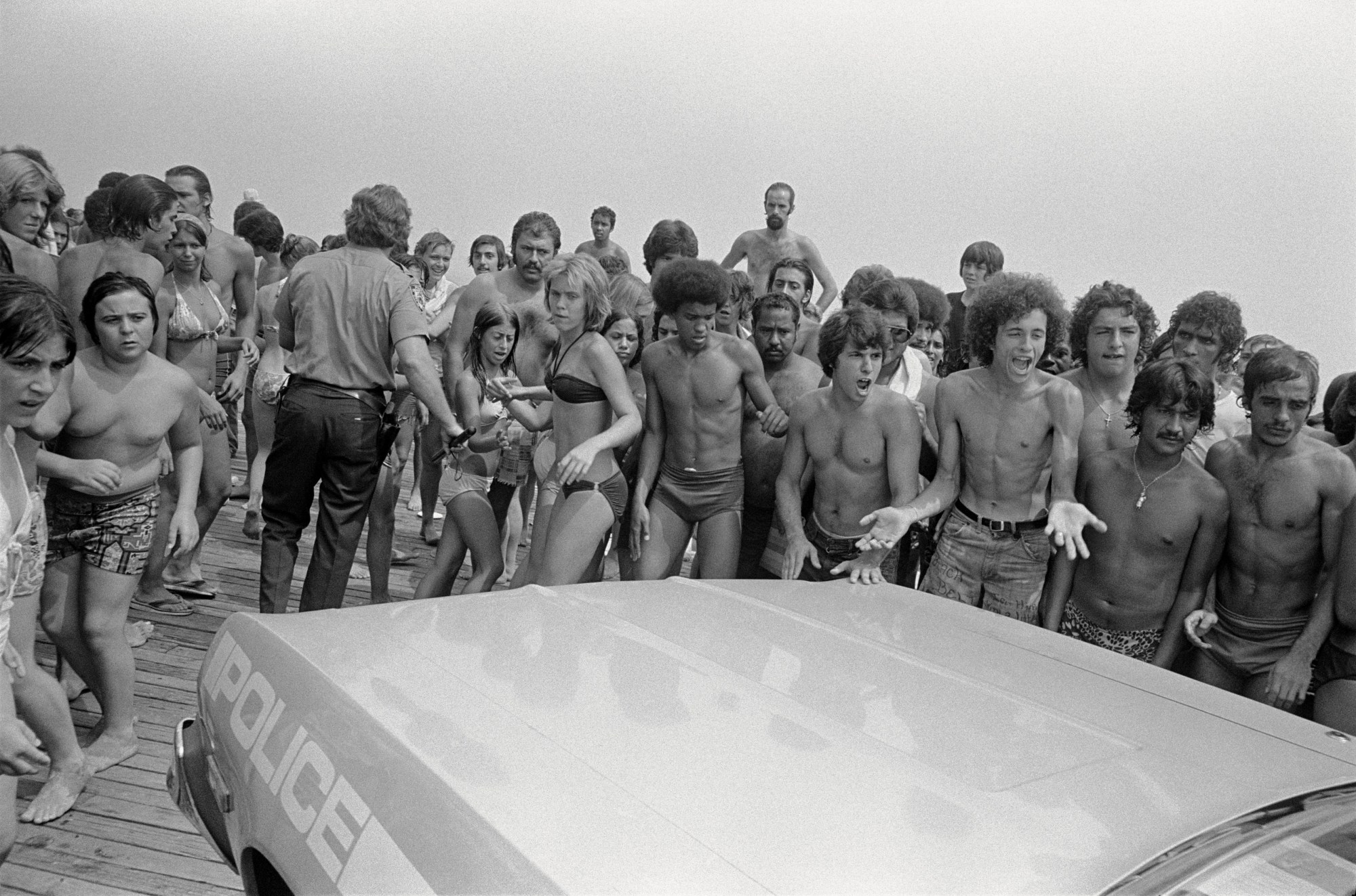 a group of people in bathing suits surrounding a police car in the 70s