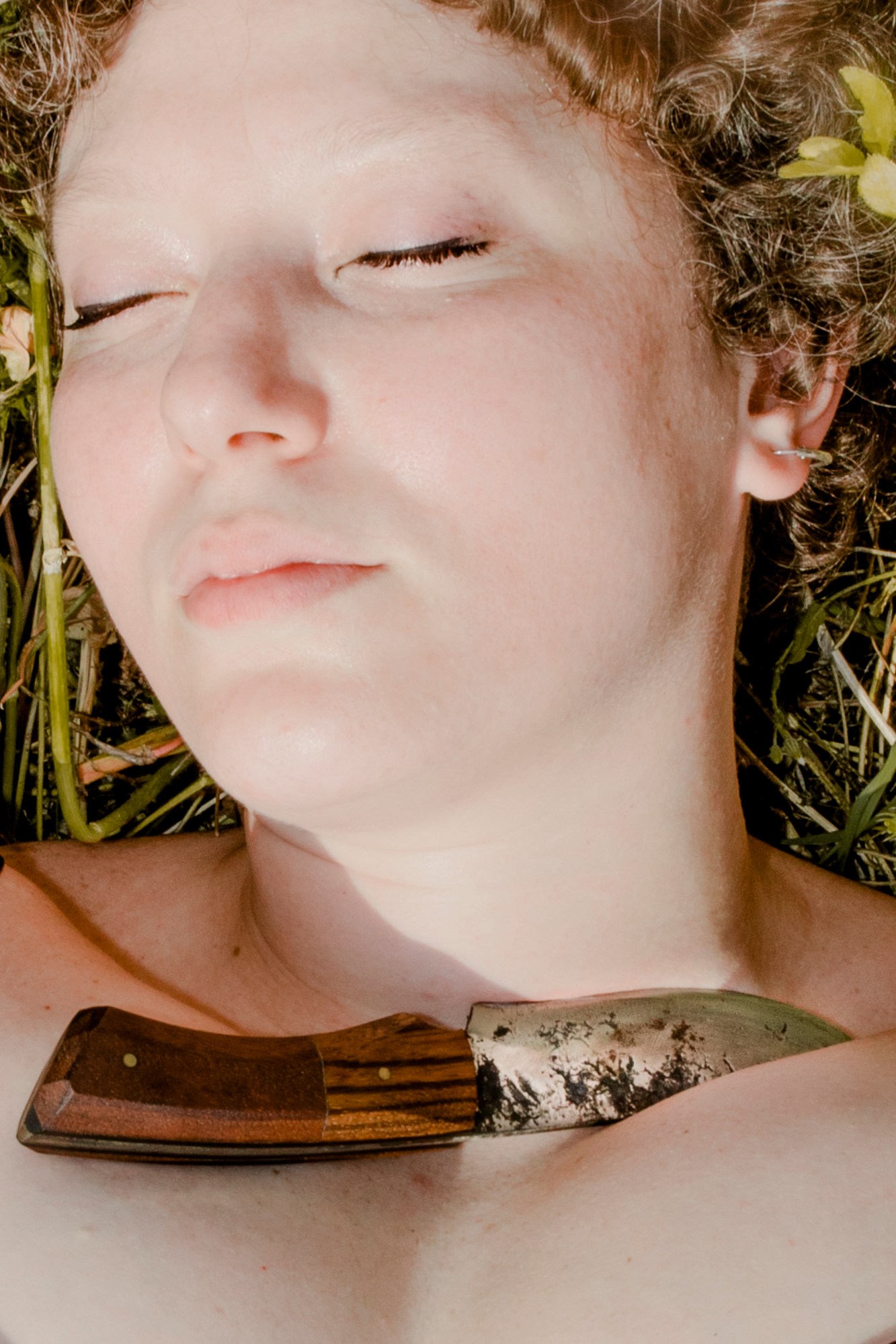 A woman lying in grass with her eyes closed with a small knife placed near her neck.