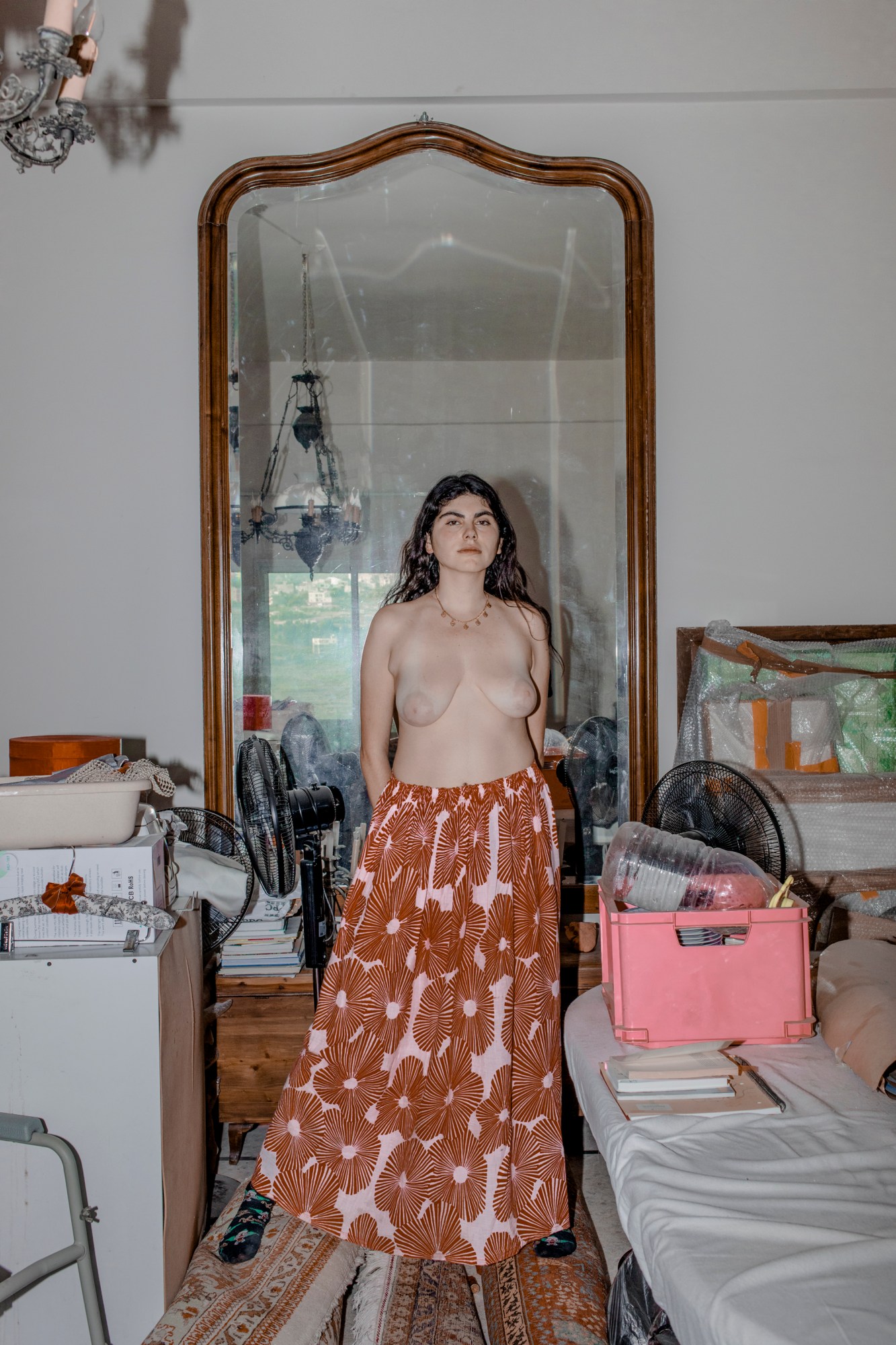 A topless woman standing near a bed and a pink filing box wearing a pink and orange floor lenth skirt.