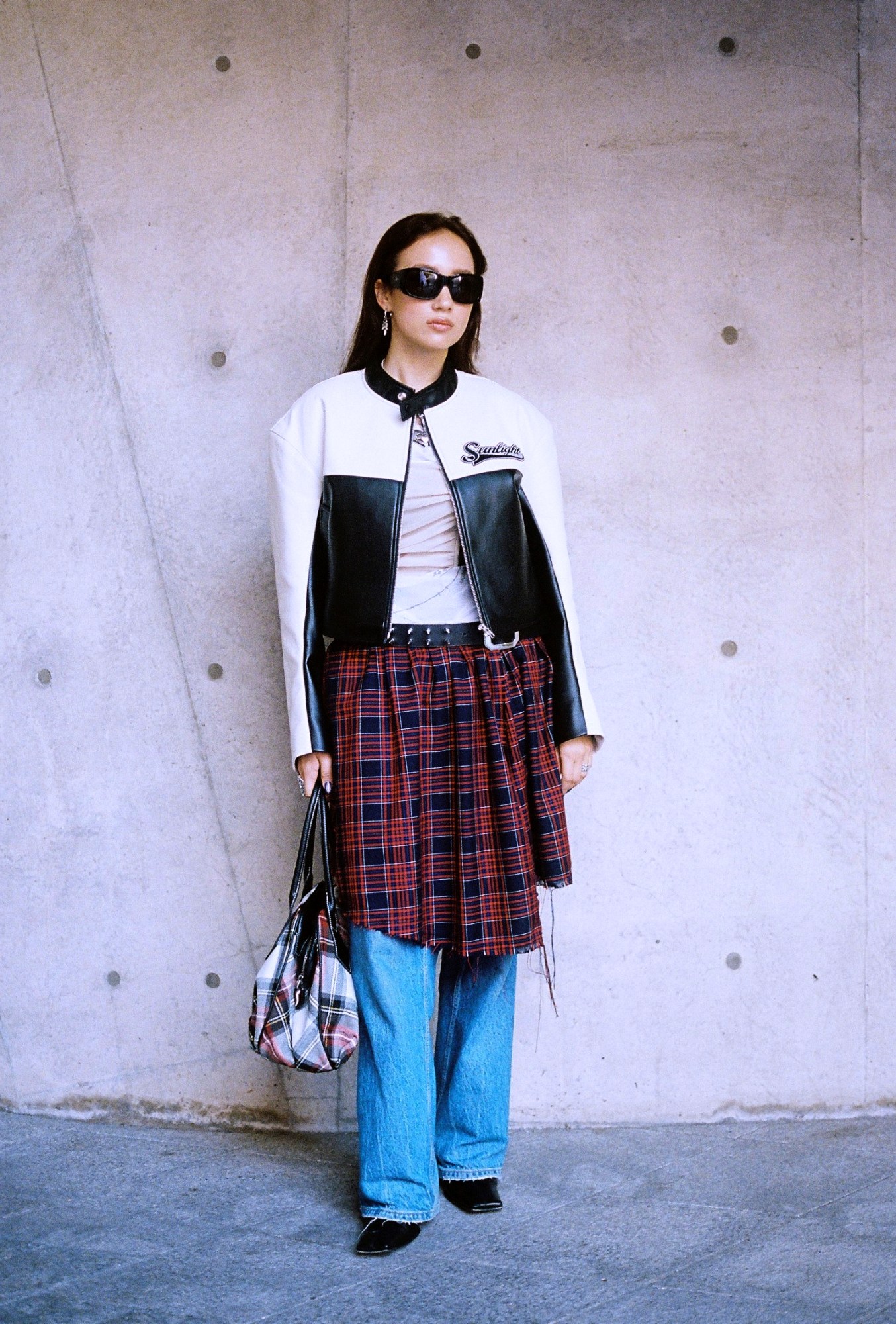 a woman wearing a kilt over jeans with a leather jacket poses against a concrete wall