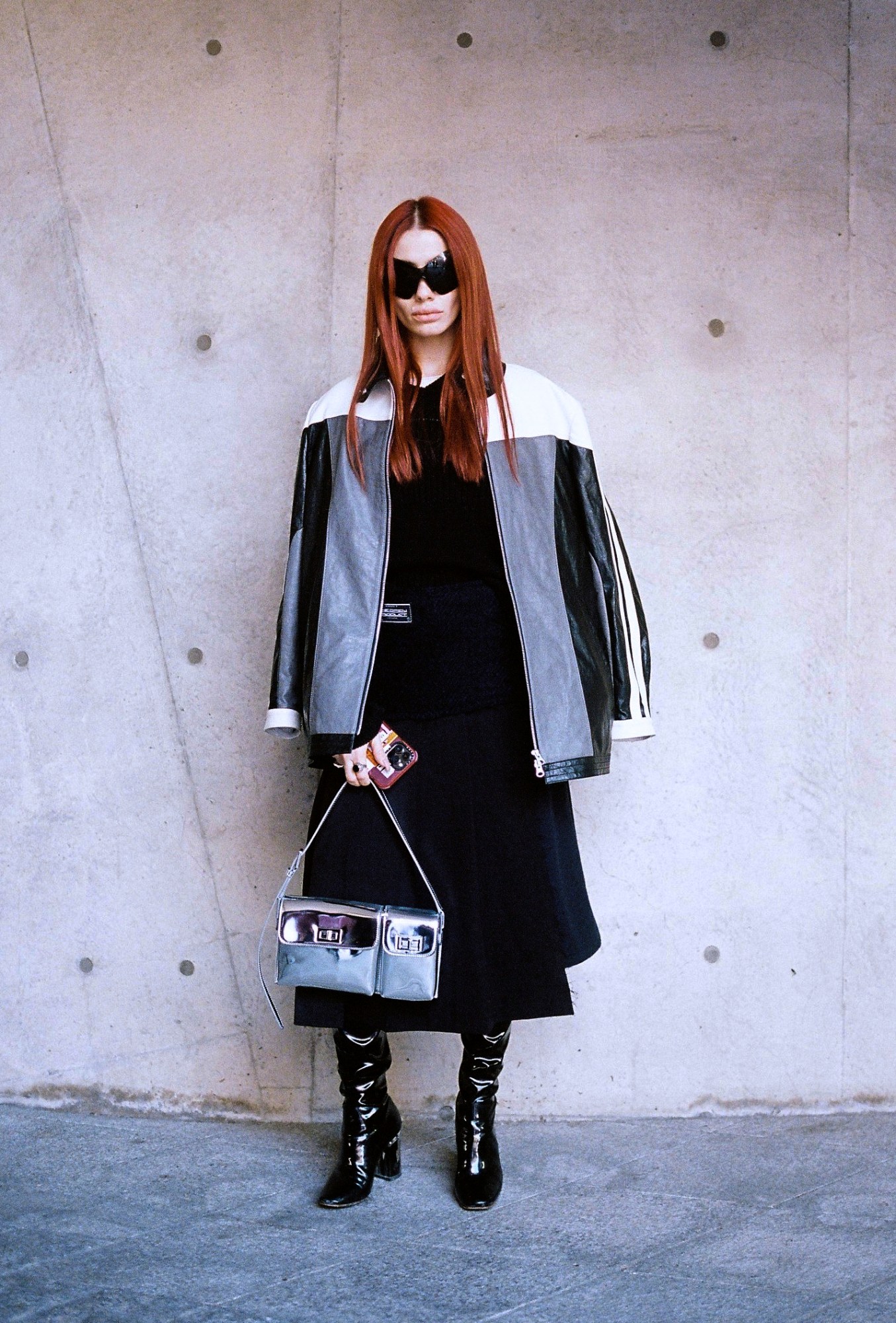 a woman with bright red hair and large sunglasses poses with a leather biker jacket over her shoulders