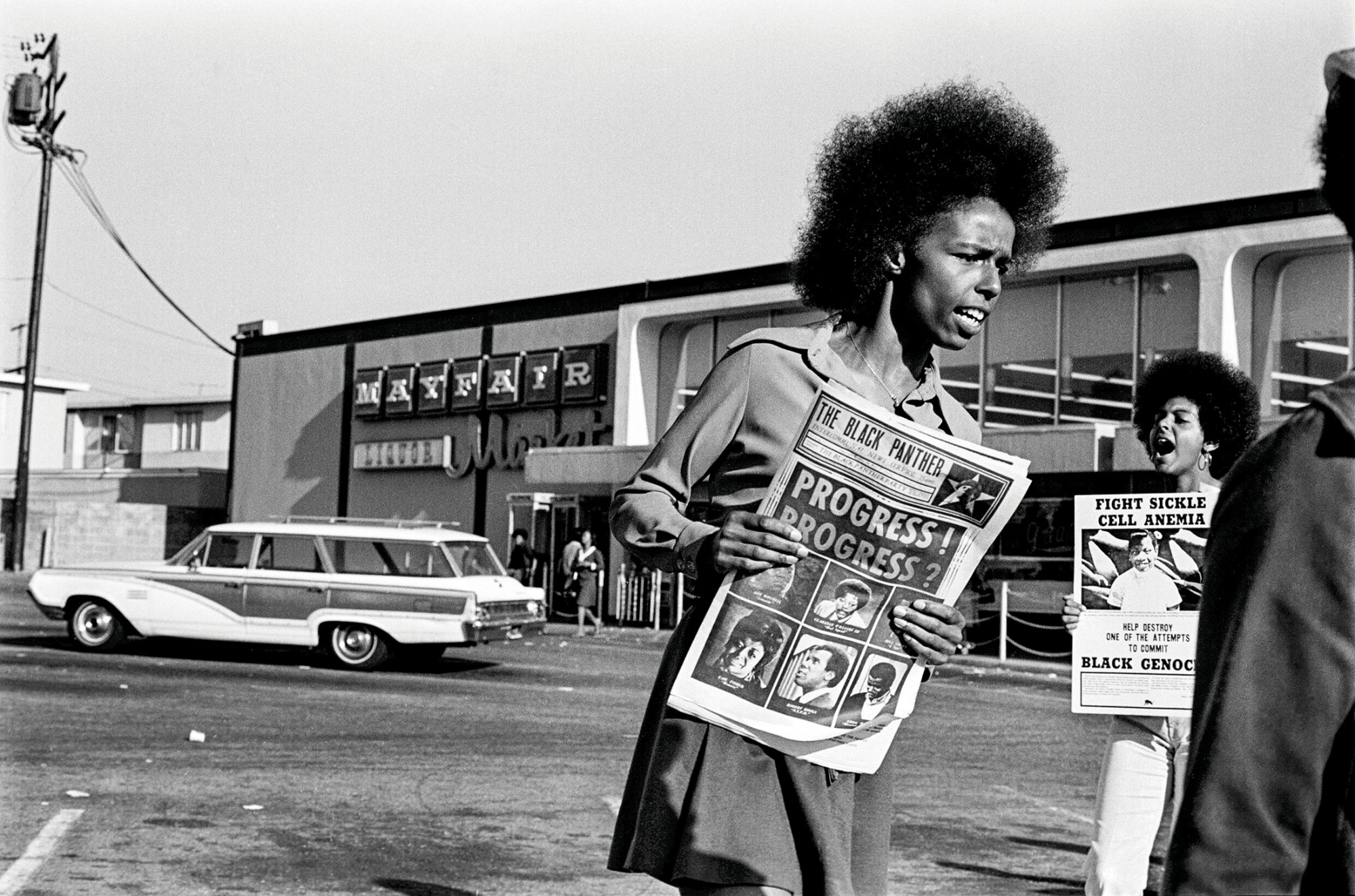 black-and-white photo of a women of the black panther party holding a poster and speaking out outside a supermarket