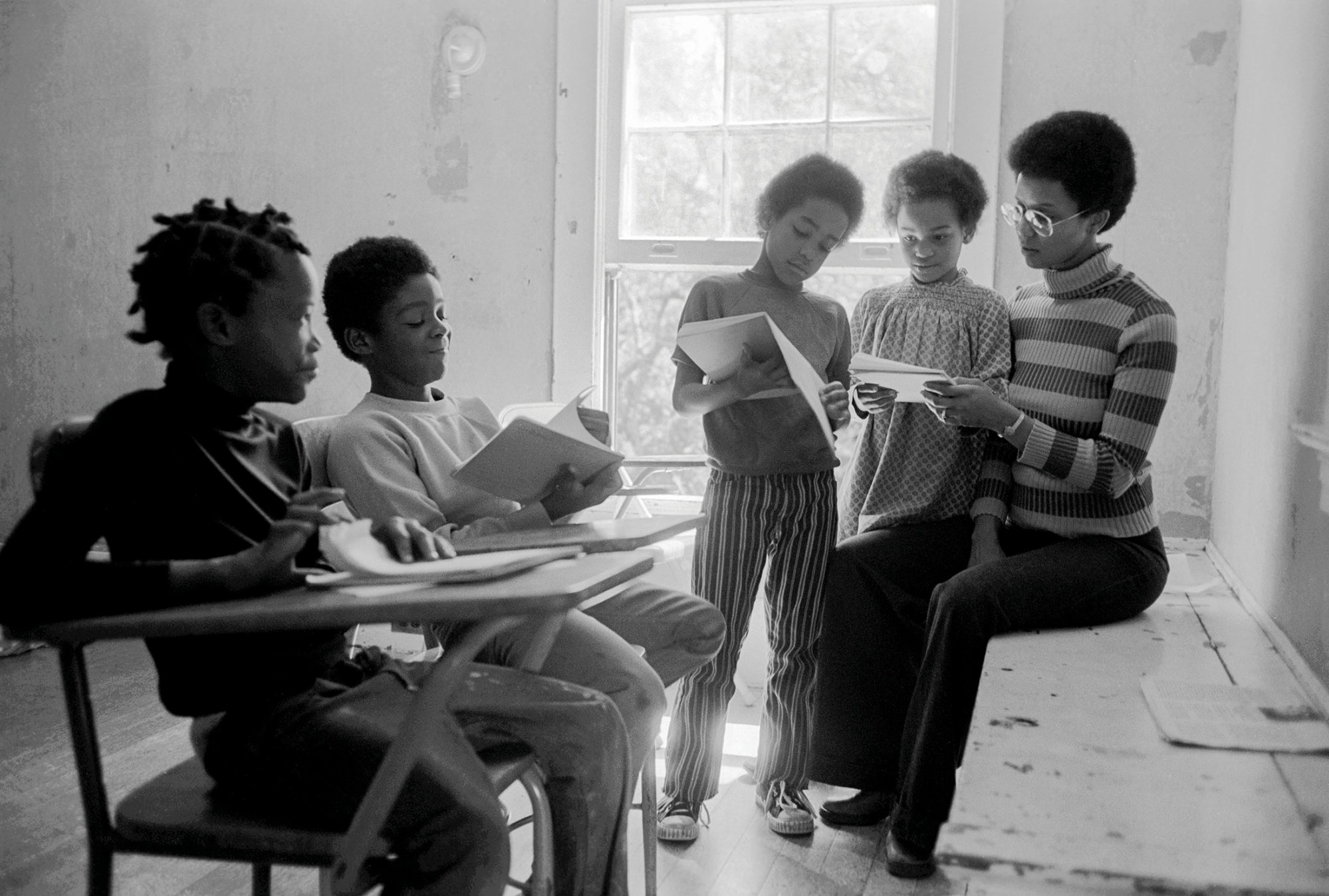 black-and-white photo of a woman of the black panther party reading with kids at a school desk