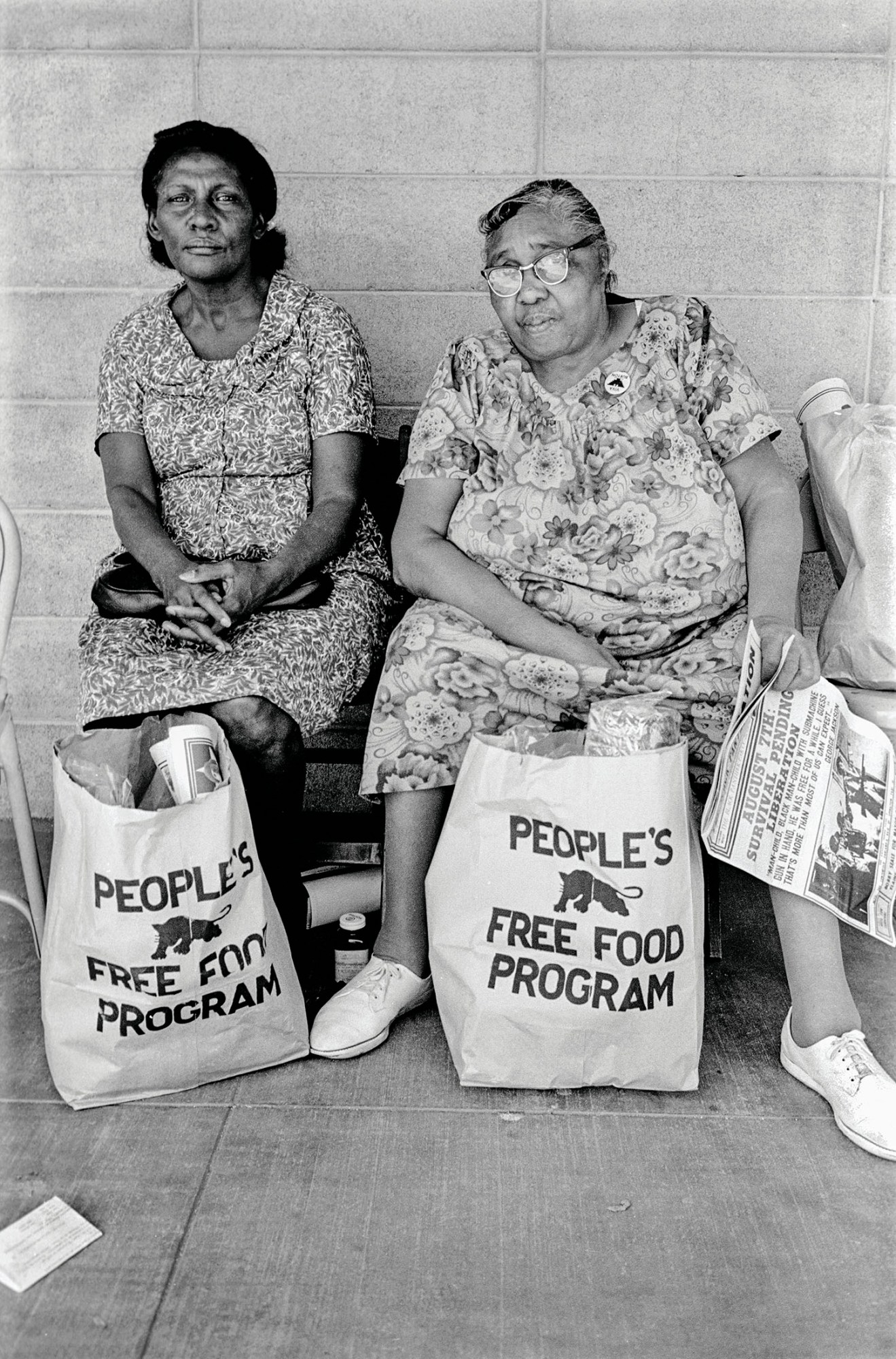 black-and-white photo of two women of the black panther party sitting on a bench with people's free food program bags filled with foods