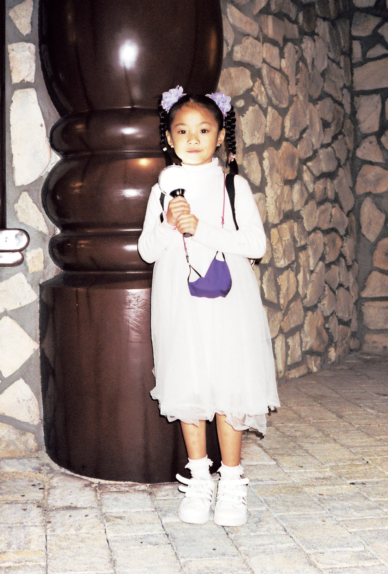 a young girl in a white dress and high pigtails clutches a light toy, a face mask hanging around her neck