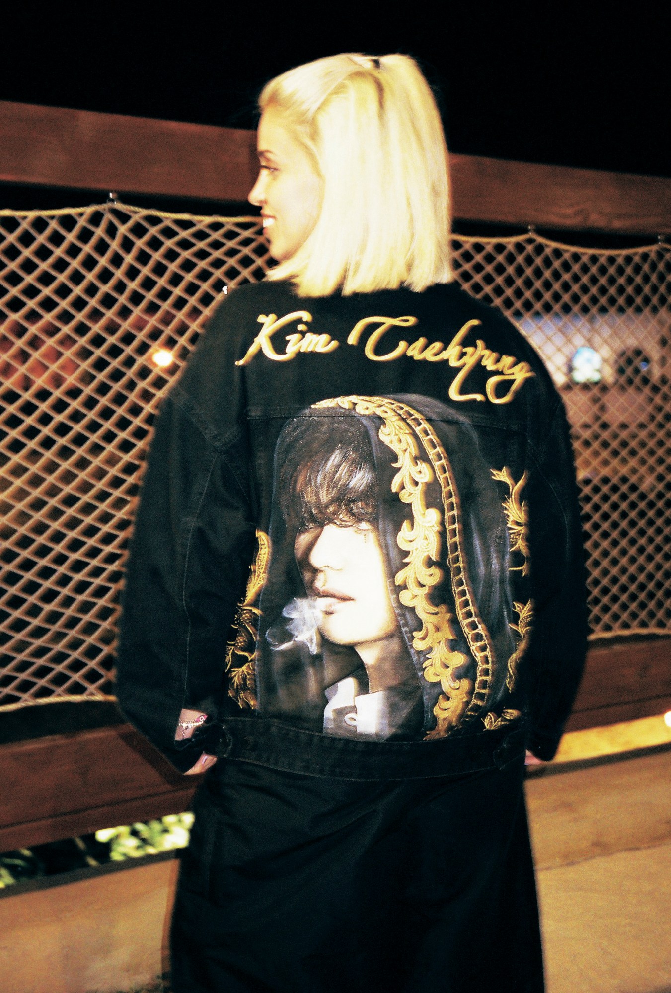 a blonde model turns to show the back of her jacket, featuring a gold embossed portrait of kim taehyung