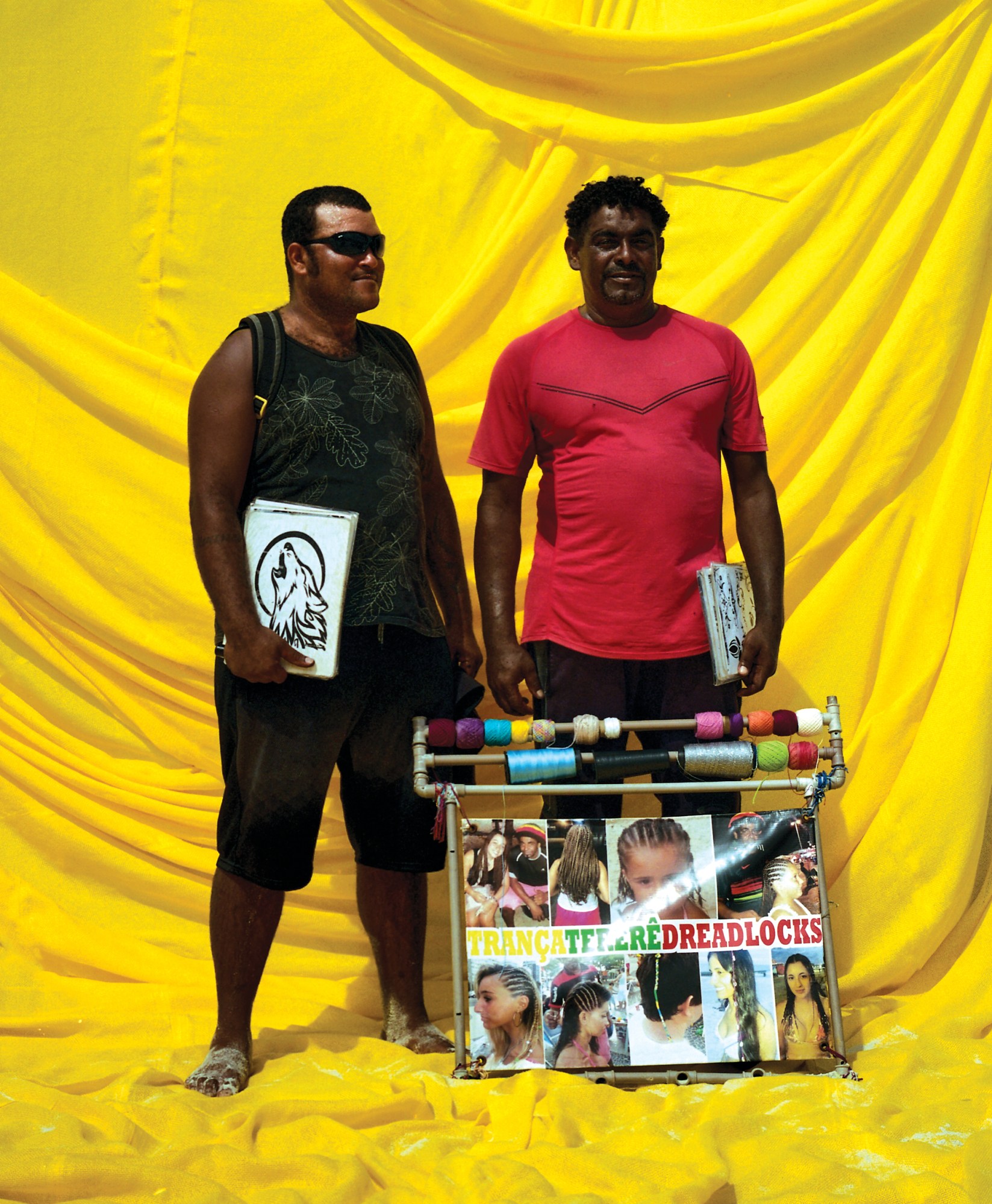 two brazilian beach sellers with a sign for braids against a yellow backdrop