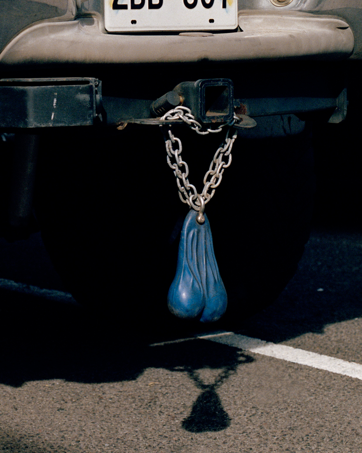 Photograph by Caroline Tompkins of a chain with testicle looking decoration hanging from a car number plate