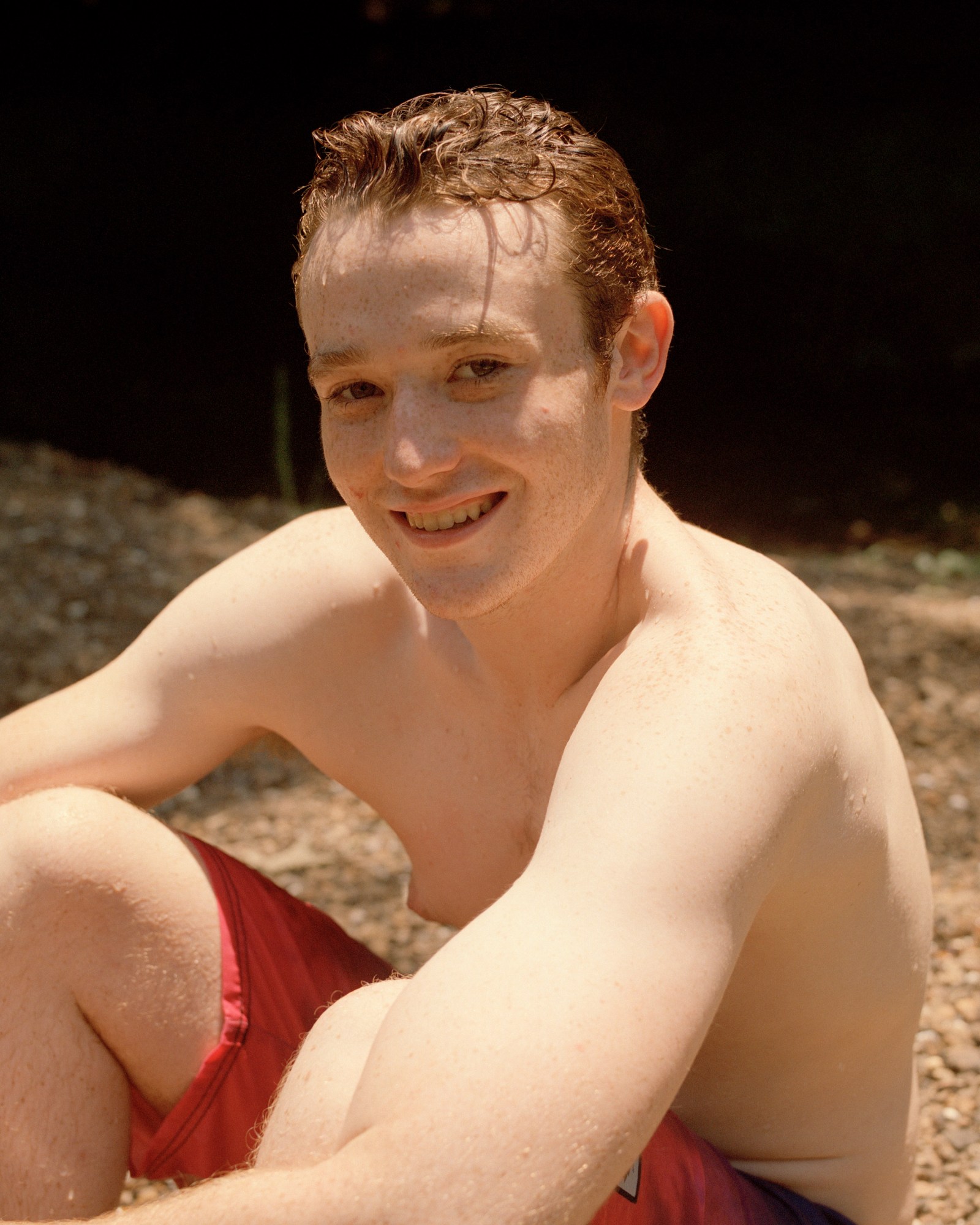 Photograph by Caroline Tompkins of a boy fresh out of the ocean and sitting on a pebble beach in swim shorts.