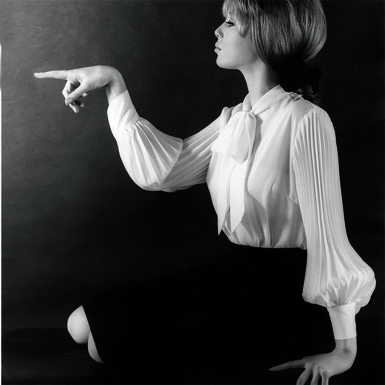 black-and-white image of pattie boyd sitting and pointing out of frame in a white shirt and black skirt