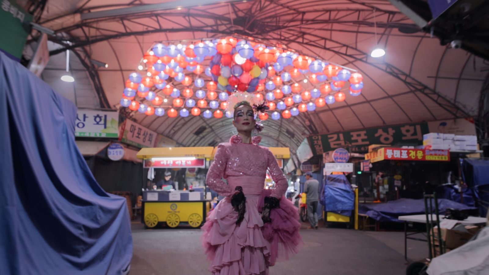 screencap from I am More, a drag queen standing in front of a well light street market