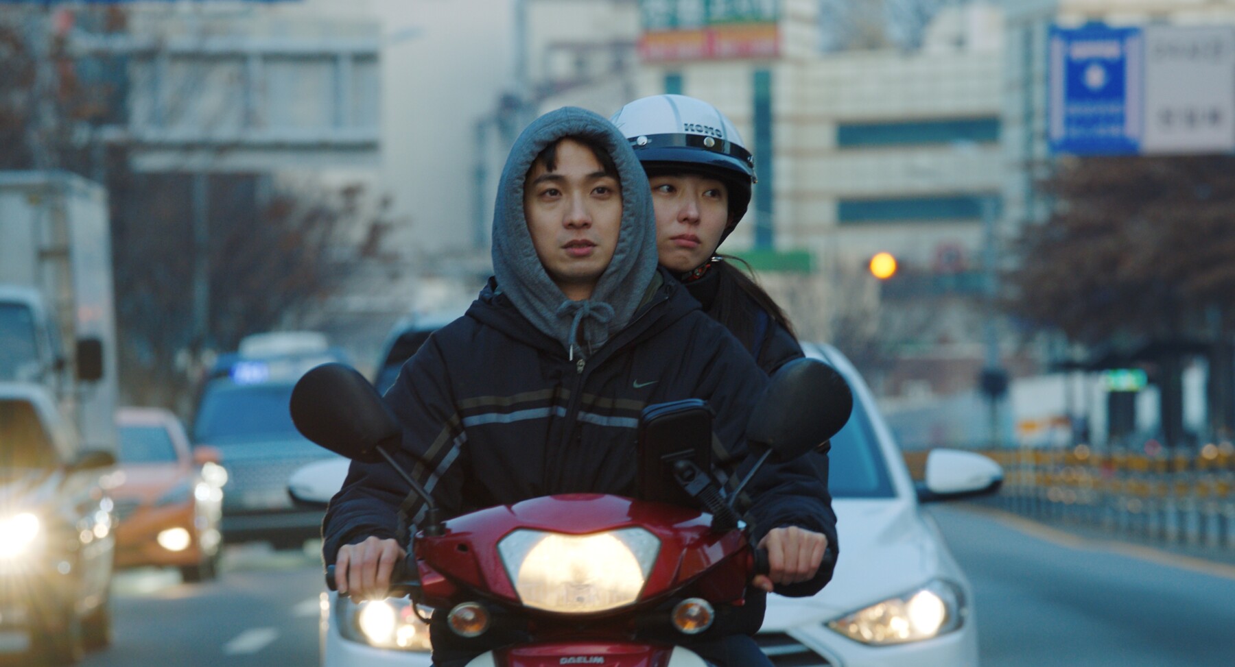 screencap from through my midwinter, two people on a red moped in front of a white car on a road